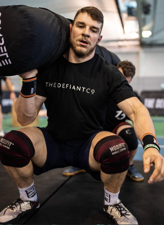 A guy wearing a The Defiant Co standard fit t-shirt whilst performing worm squats during a CrossFit workout at The Defiant Games.  The shirt is black and has the famous The Defiant Co logo across the centre of the chest in white. The shirt is roundneck, 100% cotton and extremely breathable.