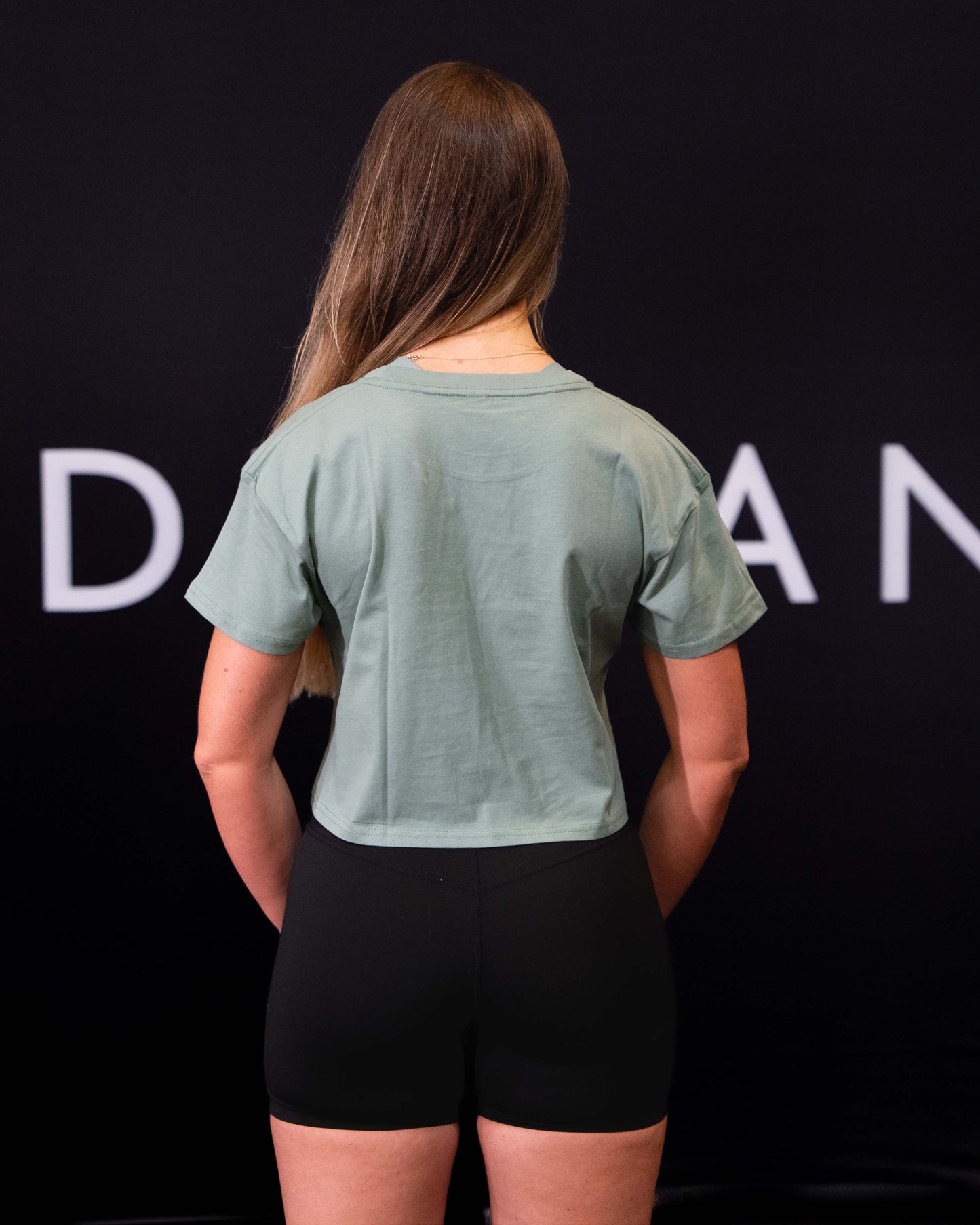A girl against a black The Defiant Co backdrop wearing the TDCo cropped t-shirt in sage green with the black DFNT. Booty shorts.