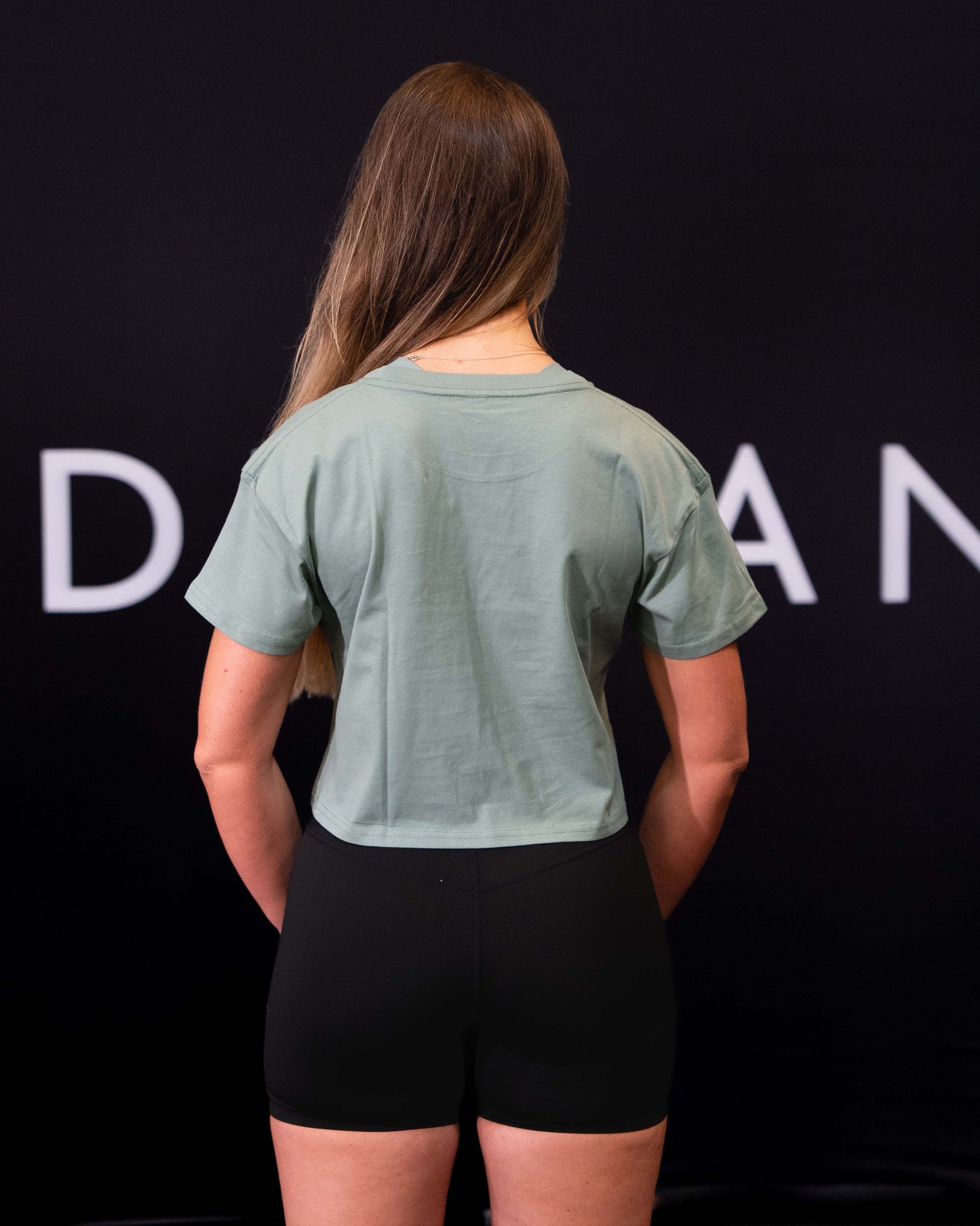 A girl against a black The Defiant Co backdrop wearing the TDCo cropped t-shirt in sage green with the black DFNT. Booty shorts.