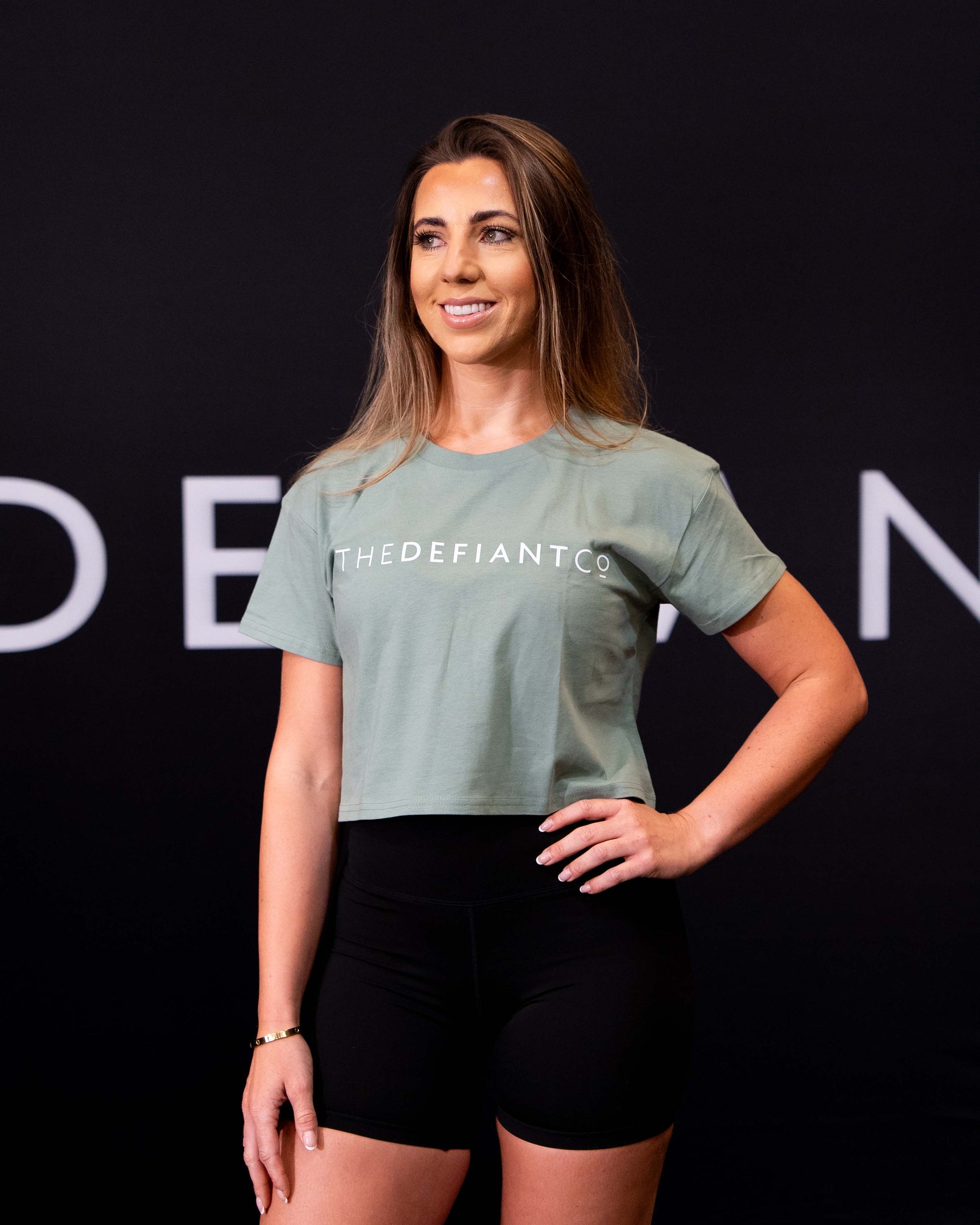 A girl against a black The Defiant Co backdrop wearing the TDCo cropped t-shirt in sage green with the black DFNT. Booty shorts.