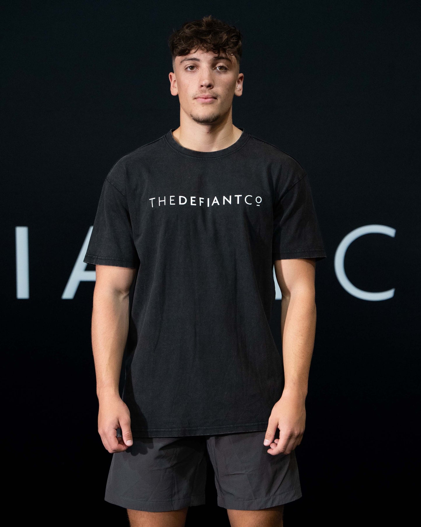 A guy against a black The Defiant Co backdrop wearing the oversized t-shirt.  The tee has the famous 'The Defiant Co' logo boldly displayed across the front. The colour is black.