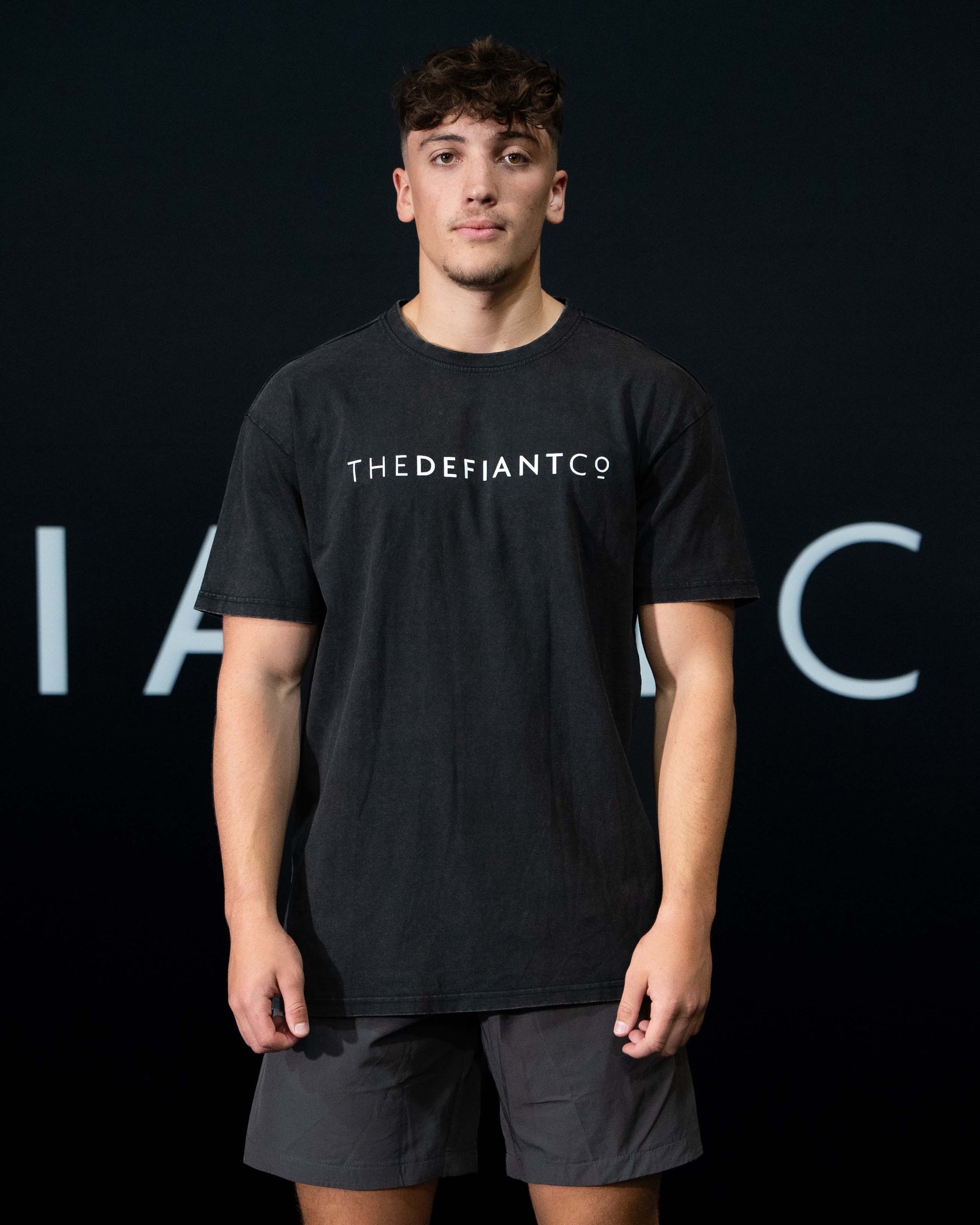 A guy against a black The Defiant Co backdrop wearing the oversized t-shirt.  The tee has the famous 'The Defiant Co' logo boldly displayed across the front. The colour is black.