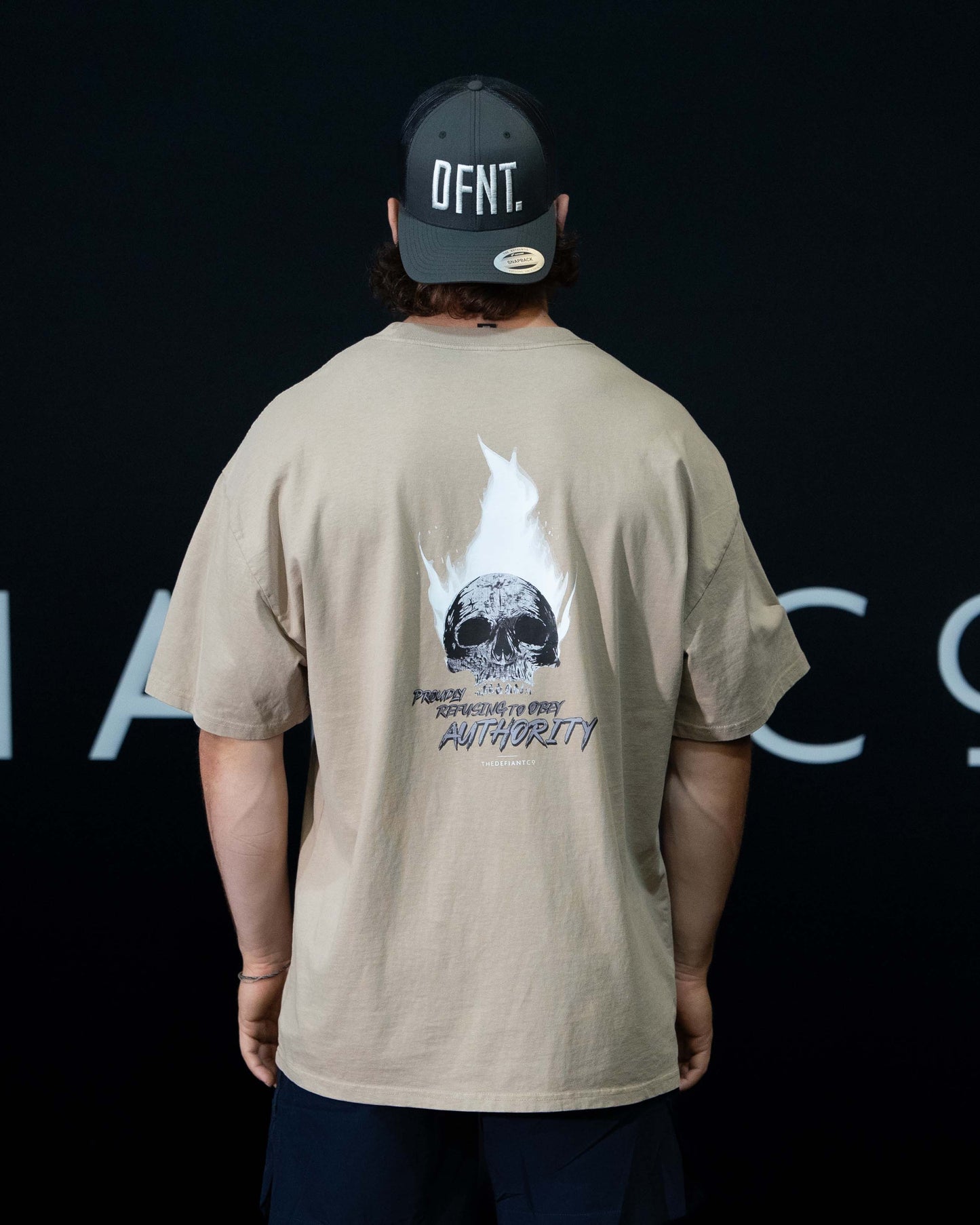 A guy against a black The Defiant Co backdrop wearing the faded bone proudly refusing to obey authority oversized tee and black performance shorts.