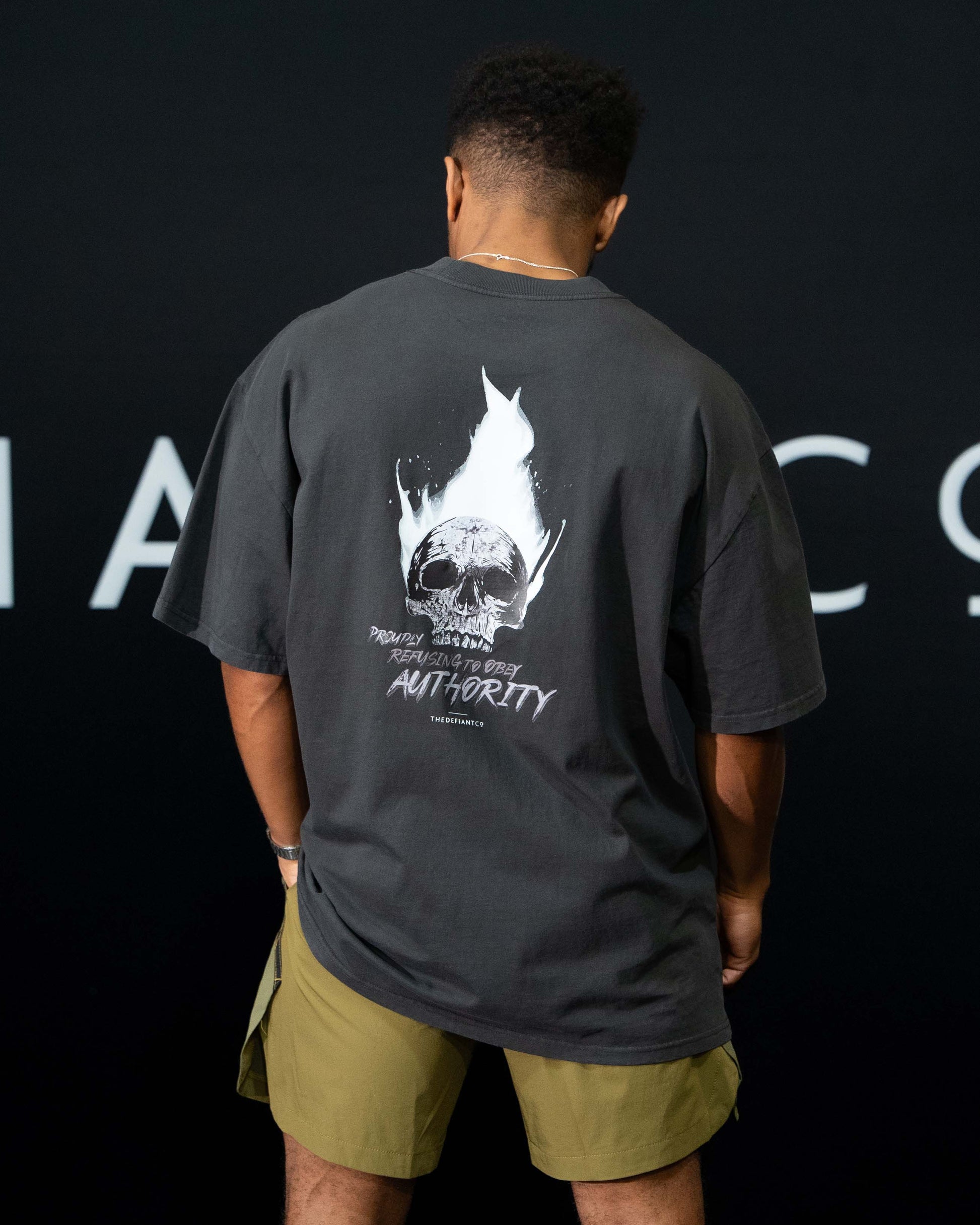A guy against a black The Defiant Co backdrop wearing the faded black proudly refusing to obey authority oversized tee and olive green performance shorts.