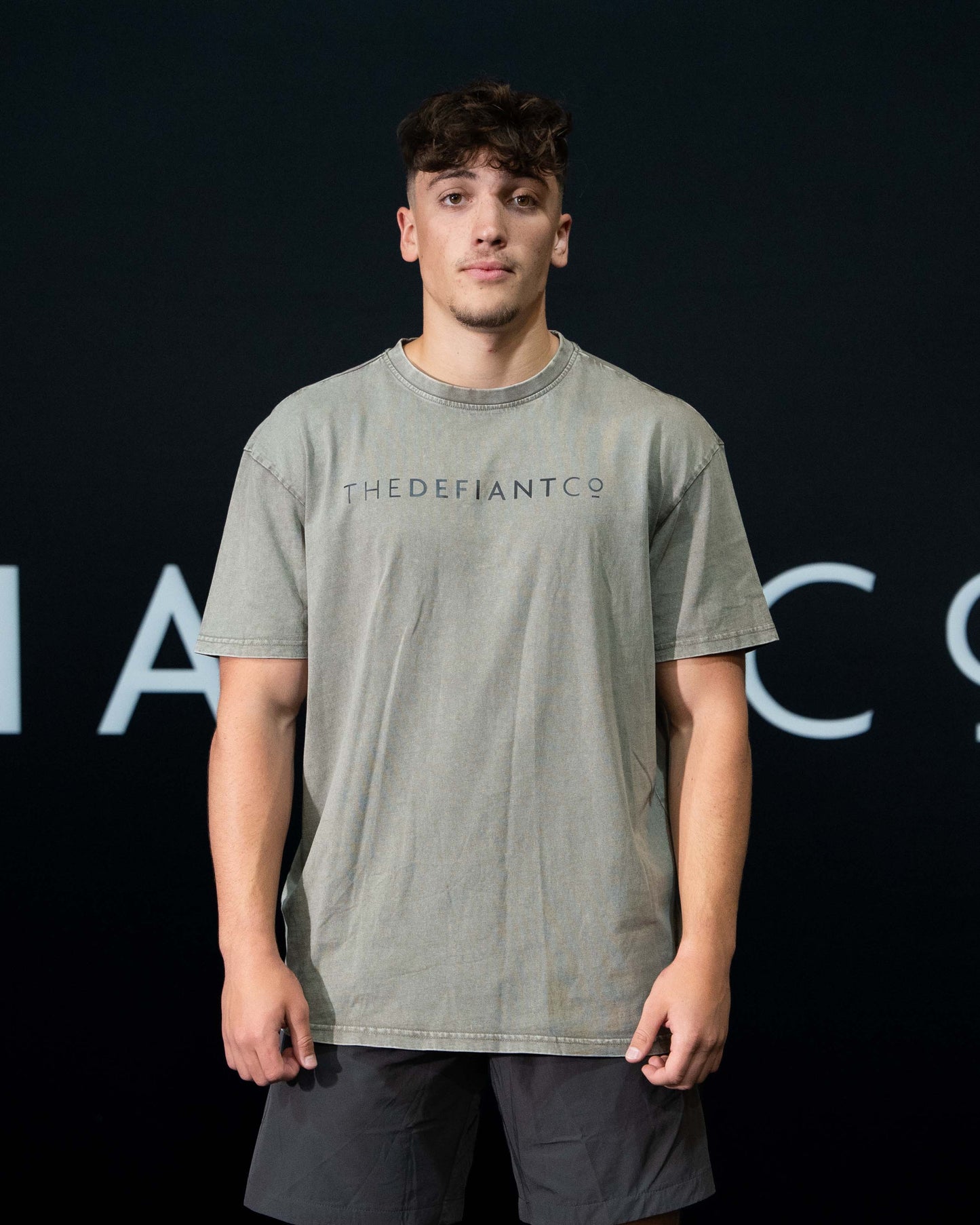 A guy against a black The Defiant Co backdrop wearing the oversized t-shirt.  The tee has the famous 'The Defiant Co' logo boldly displayed across the front. The colour is washed khaki.