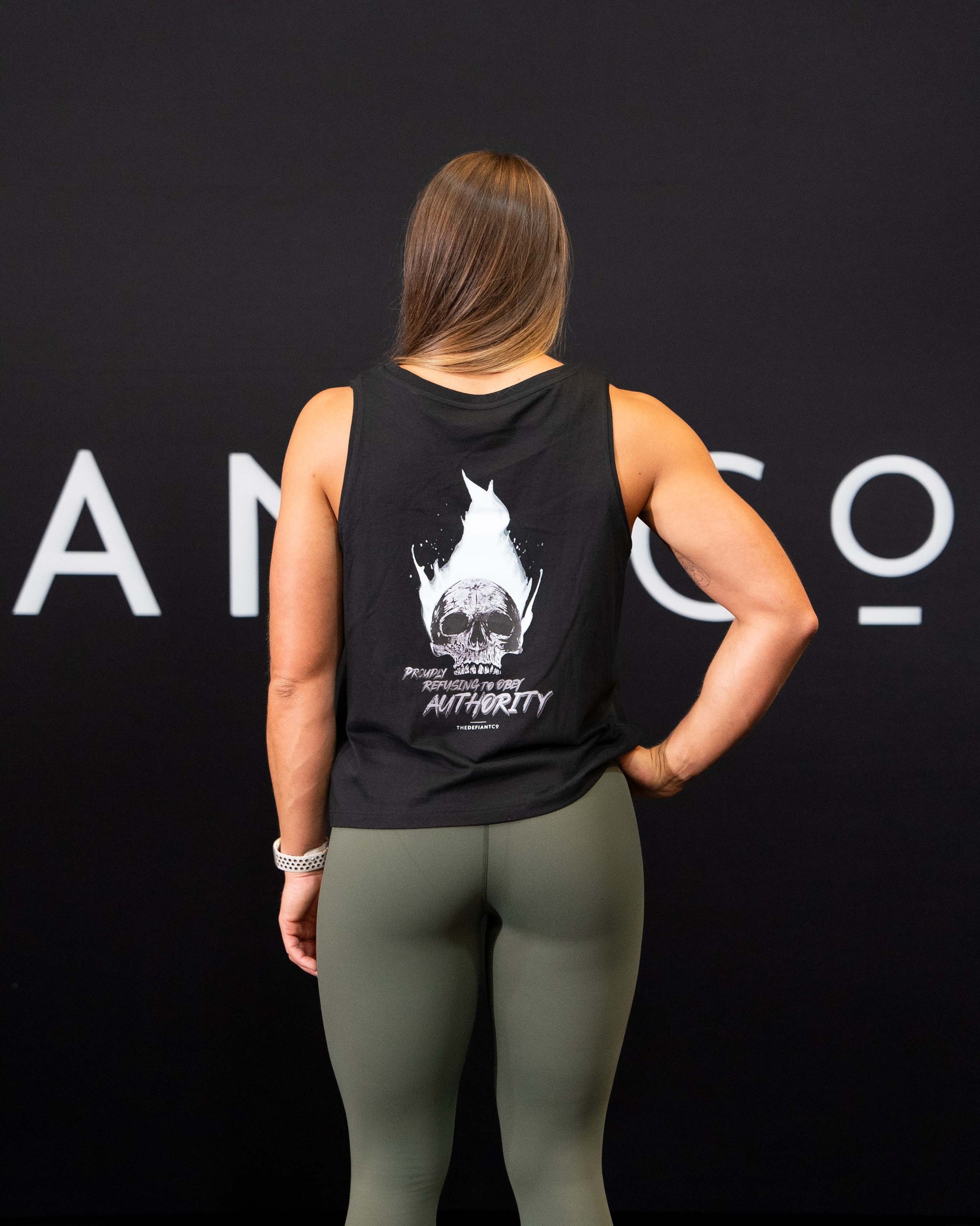 A girl against a black The Defiant Co backdrop wearing the Proudly Refusing to Obey Authority Vest Top.  The vest is slightly cropped with a high neckline.  It has a subtle left breast print and large skull and flames graphic on the back.  The colour is black.