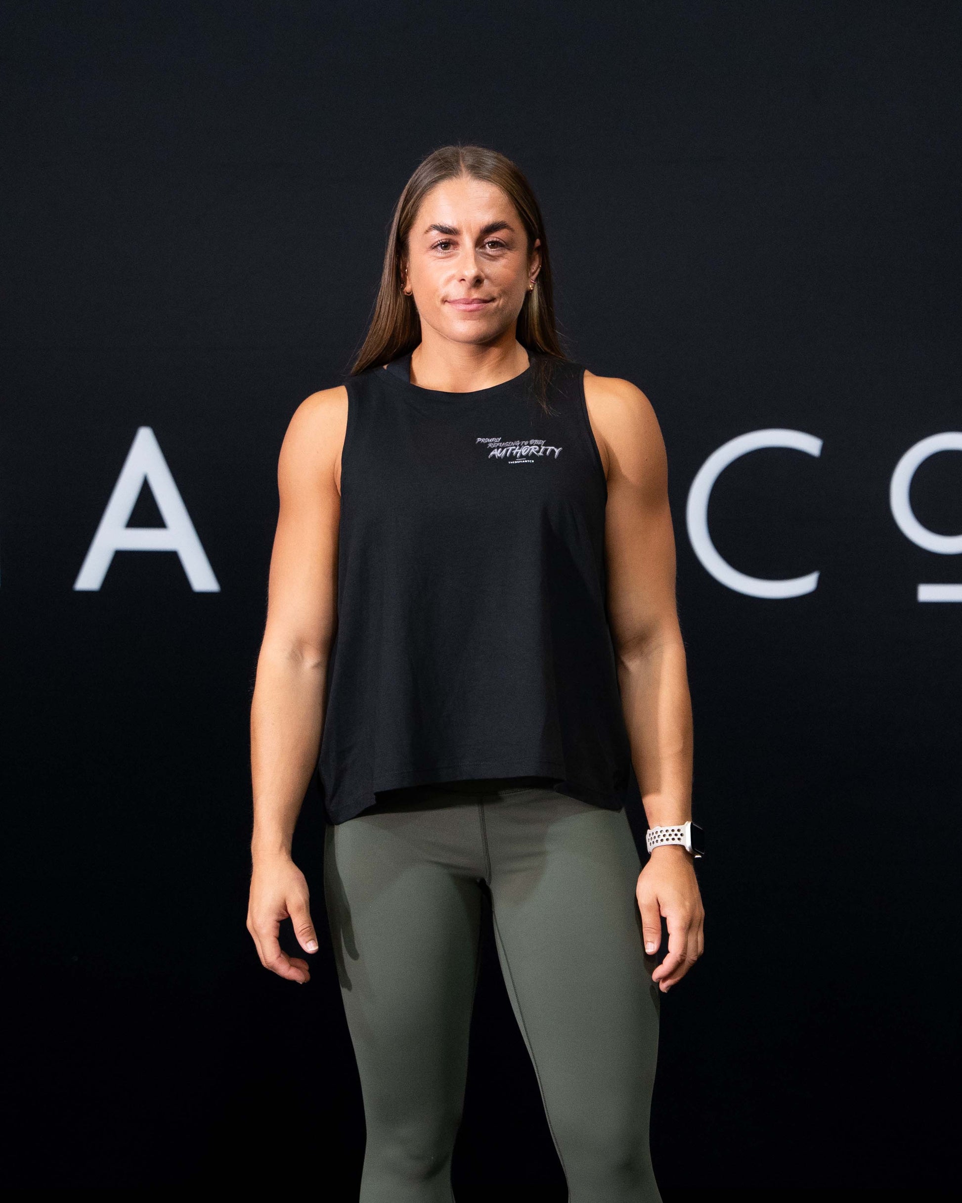 A girl against a black The Defiant Co backdrop wearing the Proudly Refusing to Obey Authority Vest Top.  The vest is slightly cropped with a high neckline.  It has a subtle left breast print and large skull and flames graphic on the back.  The colour is black.