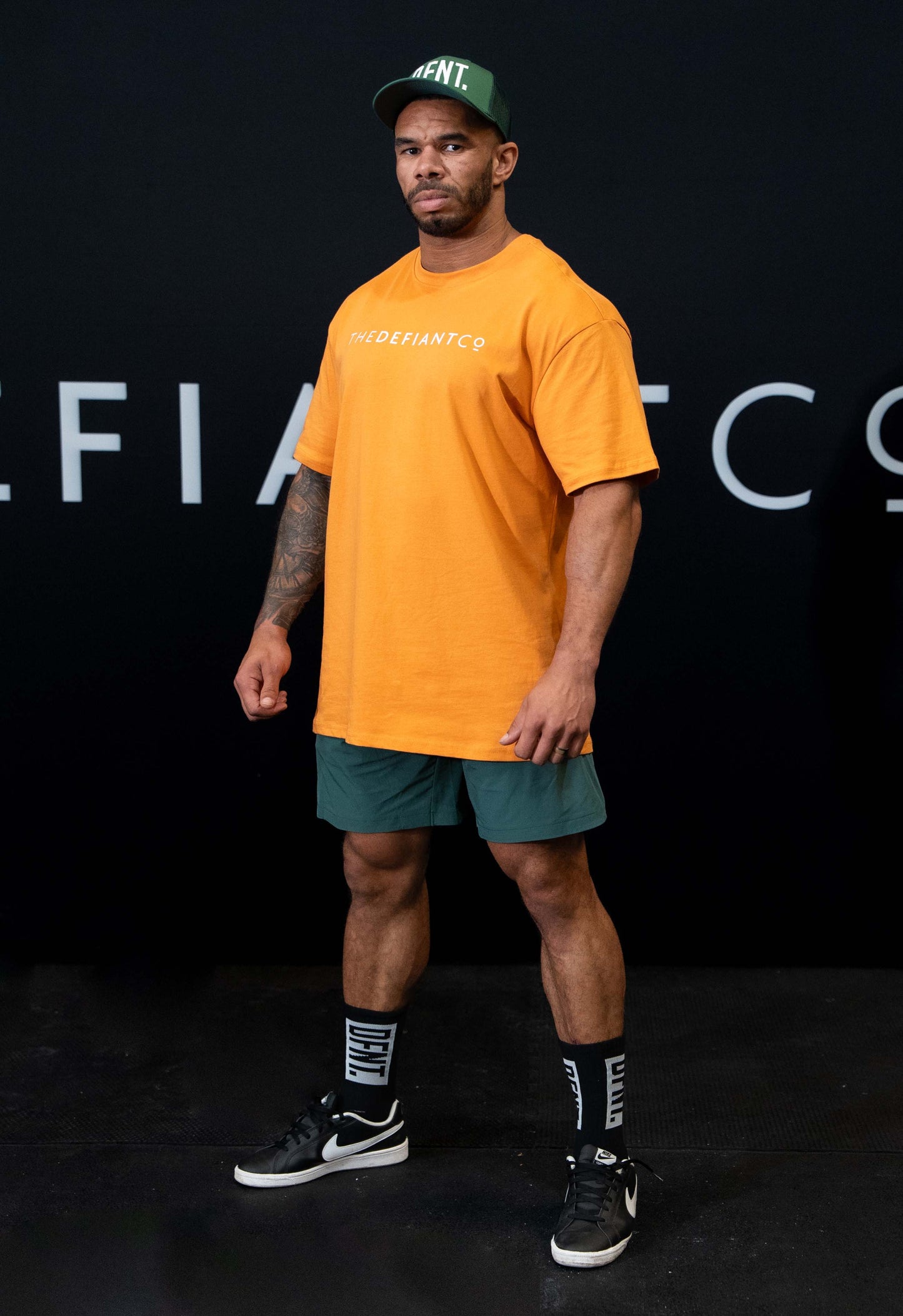 A guy against a black The Defiant Co backdrop wearing the oversized t-shirt.  The tee has the famous 'The Defiant Co' logo boldly displayed across the front. The colour is orange.