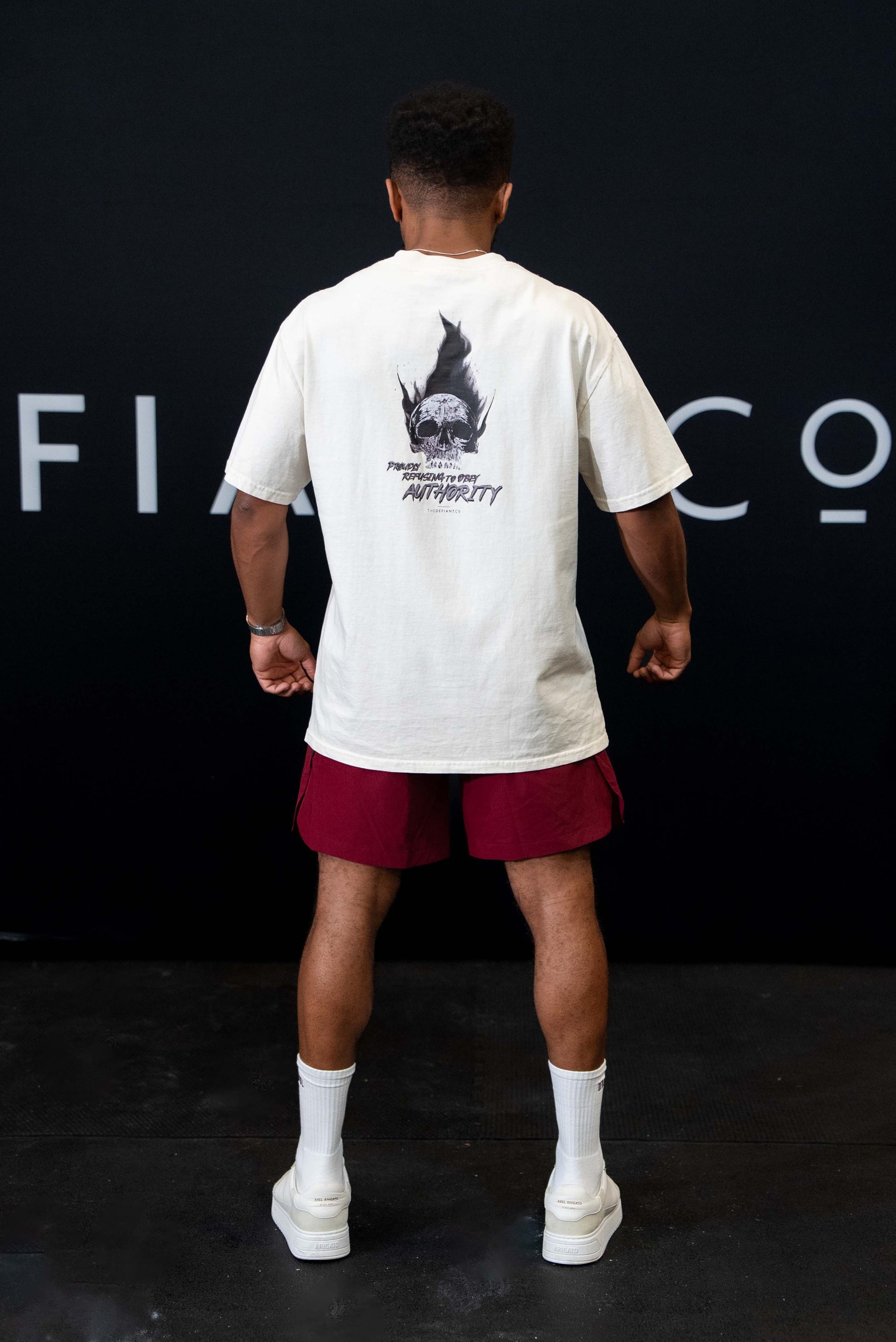 A guy against a black The Defiant Co backdrop wearing the faded ecru proudly refusing to obey authority oversized tee and burgundy performance shorts.