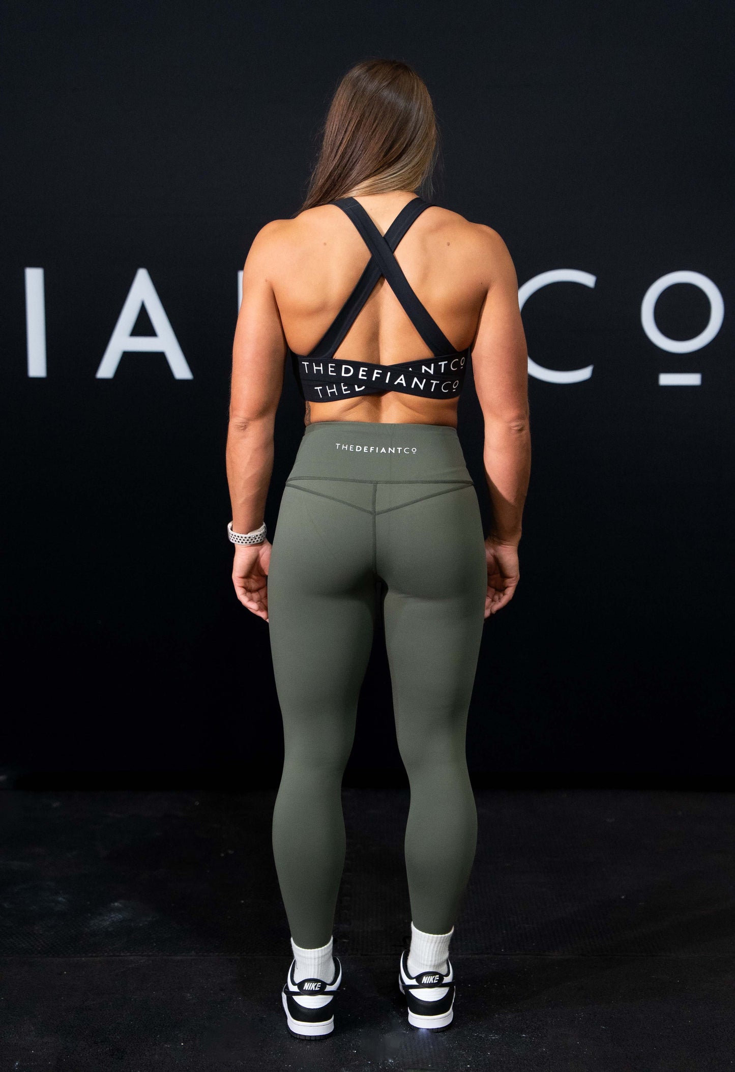 A girl against a black The Defiant Co backdrop wearing the TDCo infinity sports bra in black with the olive green performance leggings.
