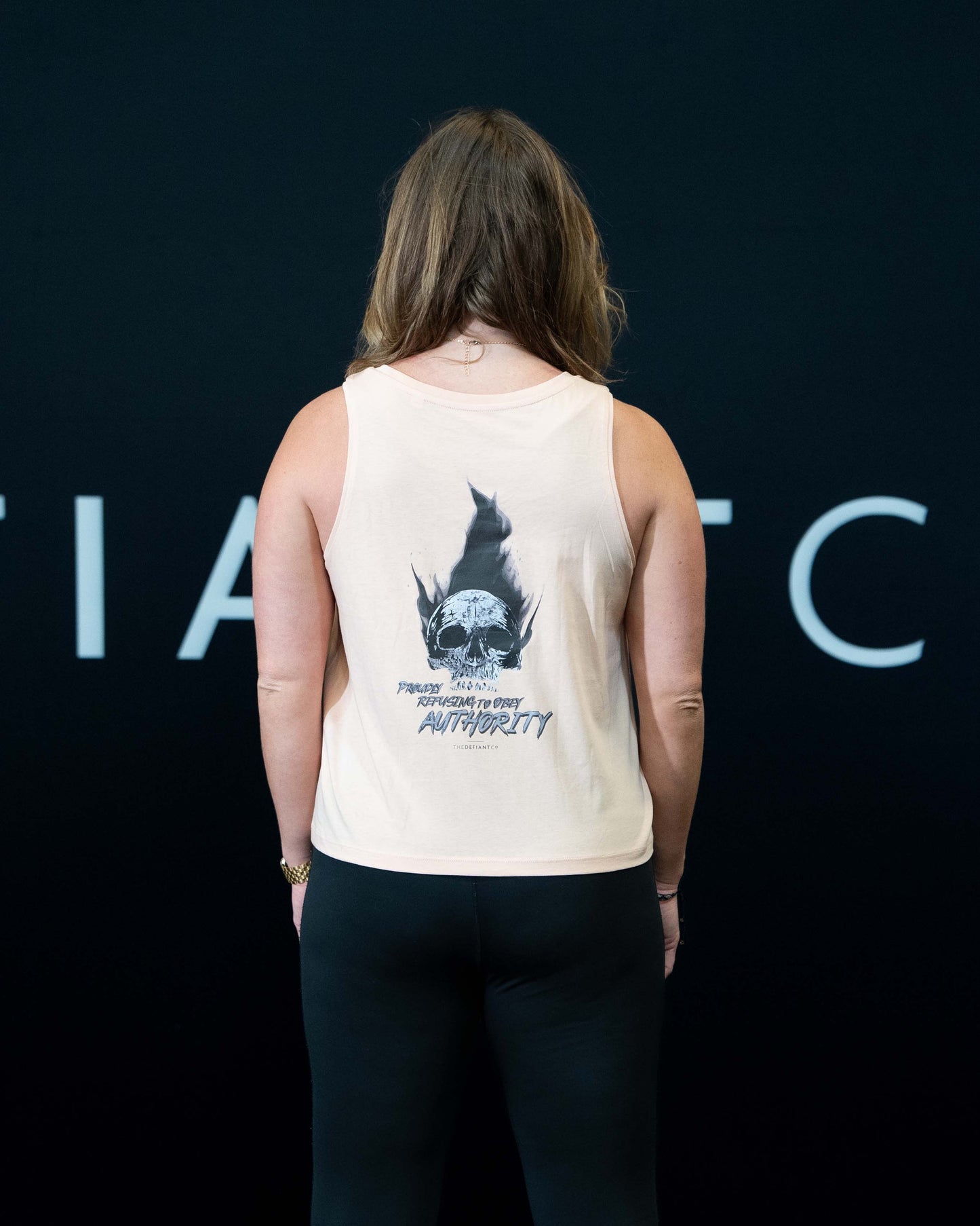 A girl against a black The Defiant Co backdrop wearing the Proudly Refusing to Obey Authority Vest Top.  The vest is slightly cropped with a high neckline.  It has a subtle left breast print and large skull and flames graphic on the back.  The colour is blush.