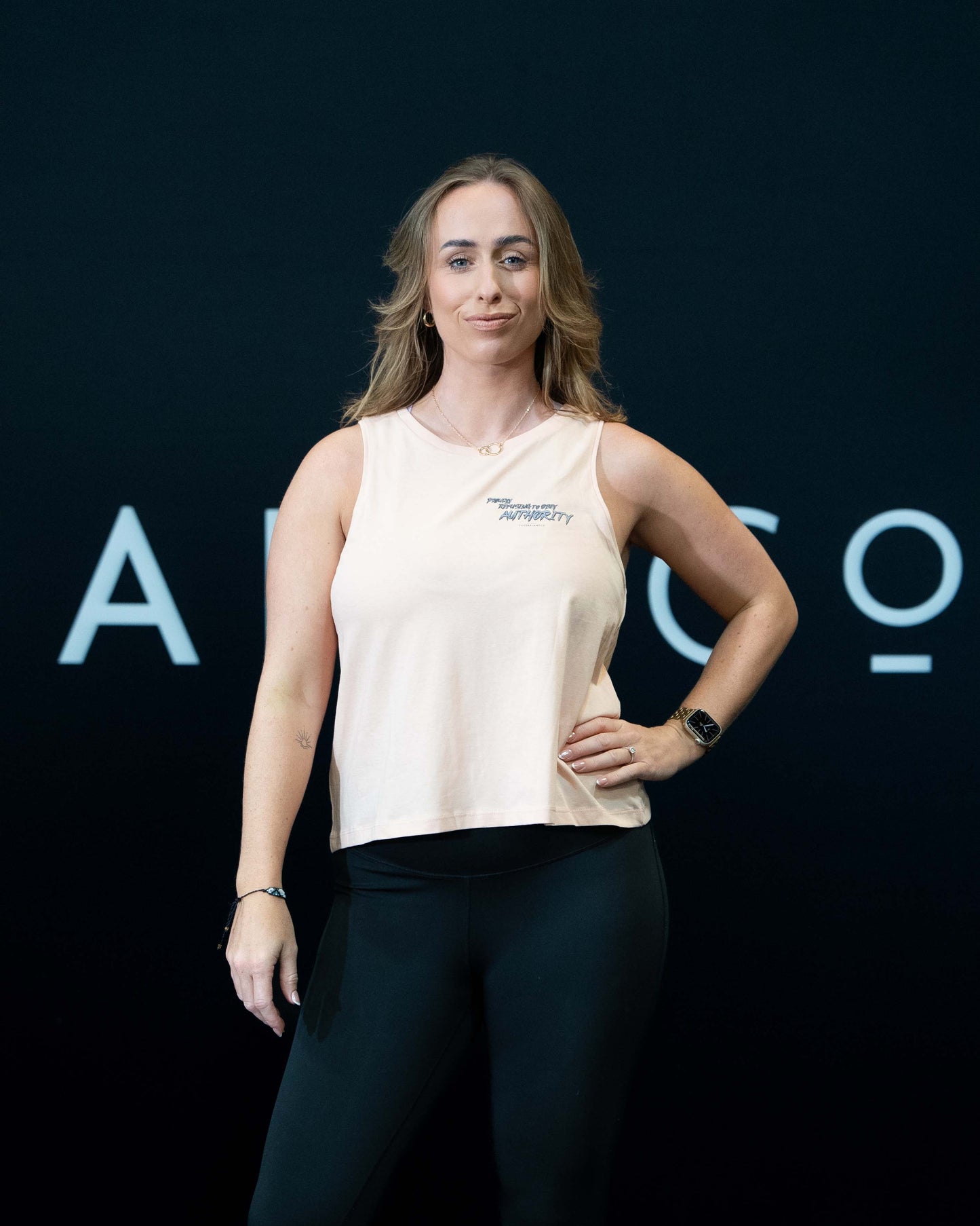 A girl against a black The Defiant Co backdrop wearing the Proudly Refusing to Obey Authority Vest Top.  The vest is slightly cropped with a high neckline.  It has a subtle left breast print and large skull and flames graphic on the back.  The colour is blush.
