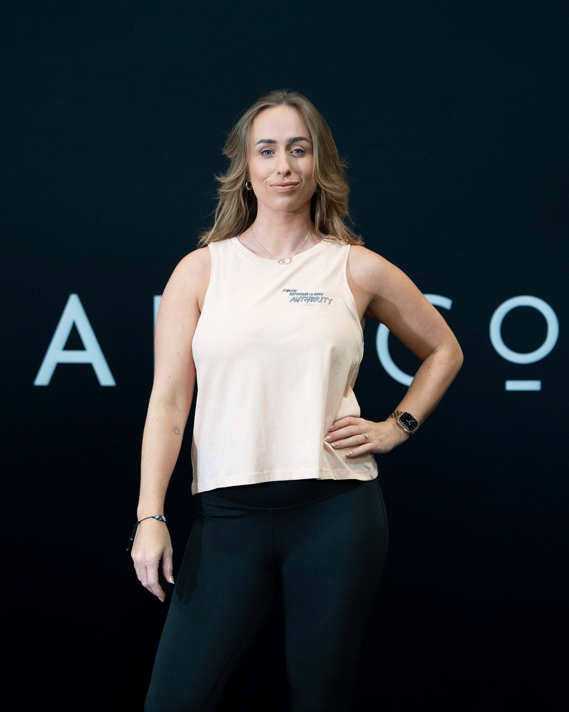 A girl against a black The Defiant Co backdrop wearing the Proudly Refusing to Obey Authority Vest Top.  The vest is slightly cropped with a high neckline.  It has a subtle left breast print and large skull and flames graphic on the back.  The colour is blush.