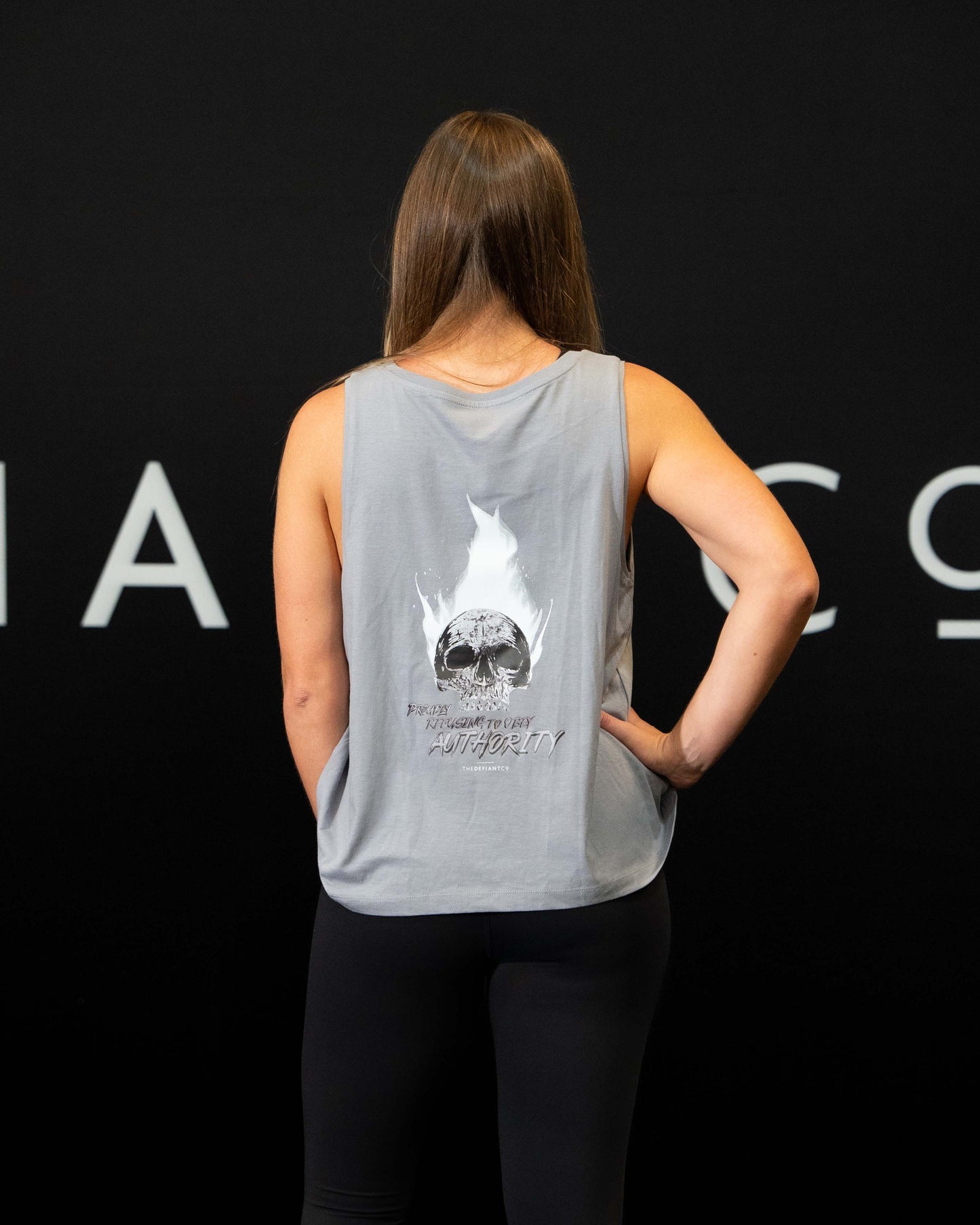 A girl against a black The Defiant Co backdrop wearing the Proudly Refusing to Obey Authority Vest Top.  The vest is slightly cropped with a high neckline.  It has a subtle left breast print and large skull and flames graphic on the back.  The colour is lava grey.