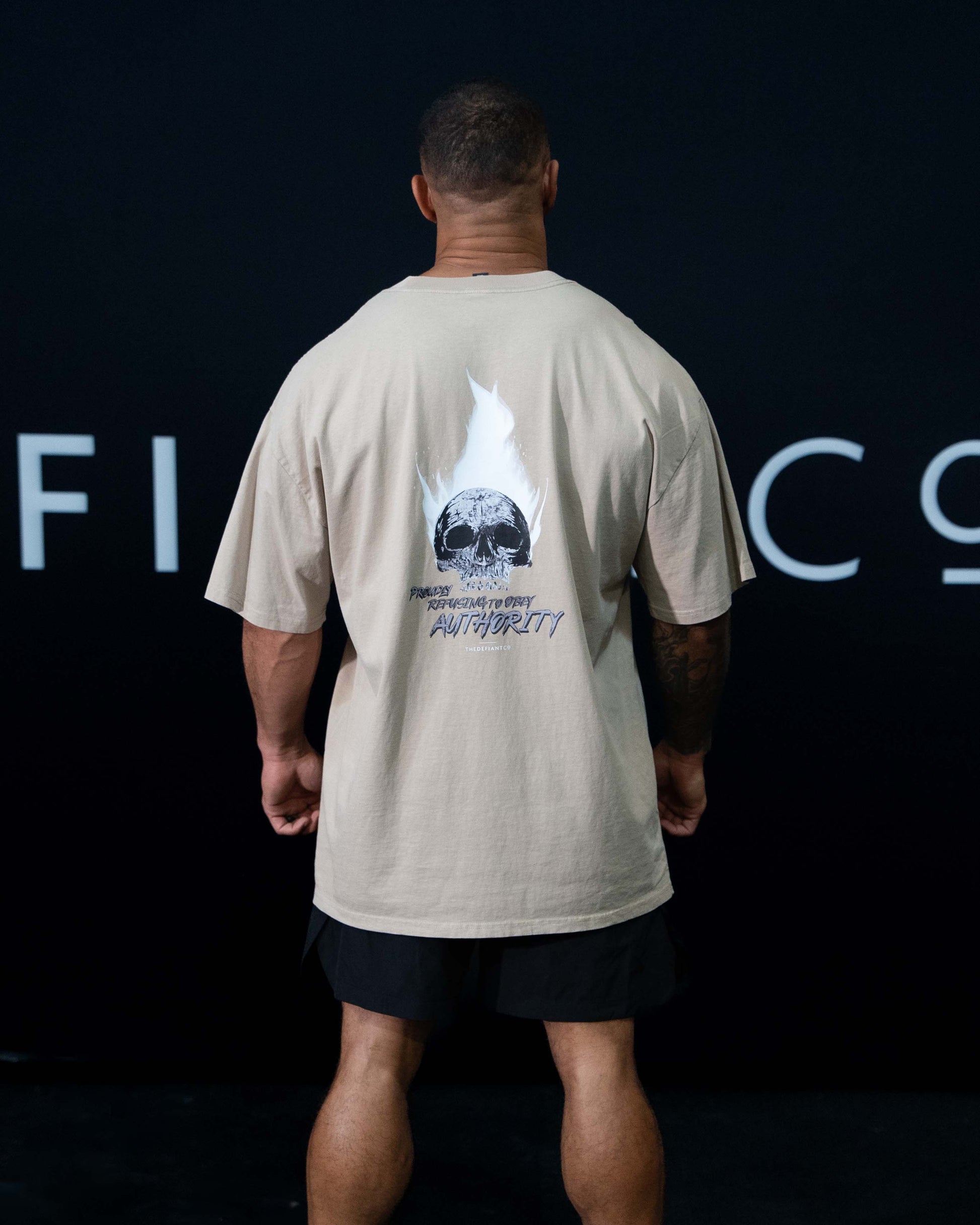 A guy against a black The Defiant Co backdrop wearing the faded khaki proudly refusing to obey authority oversized tee and black performance shorts.