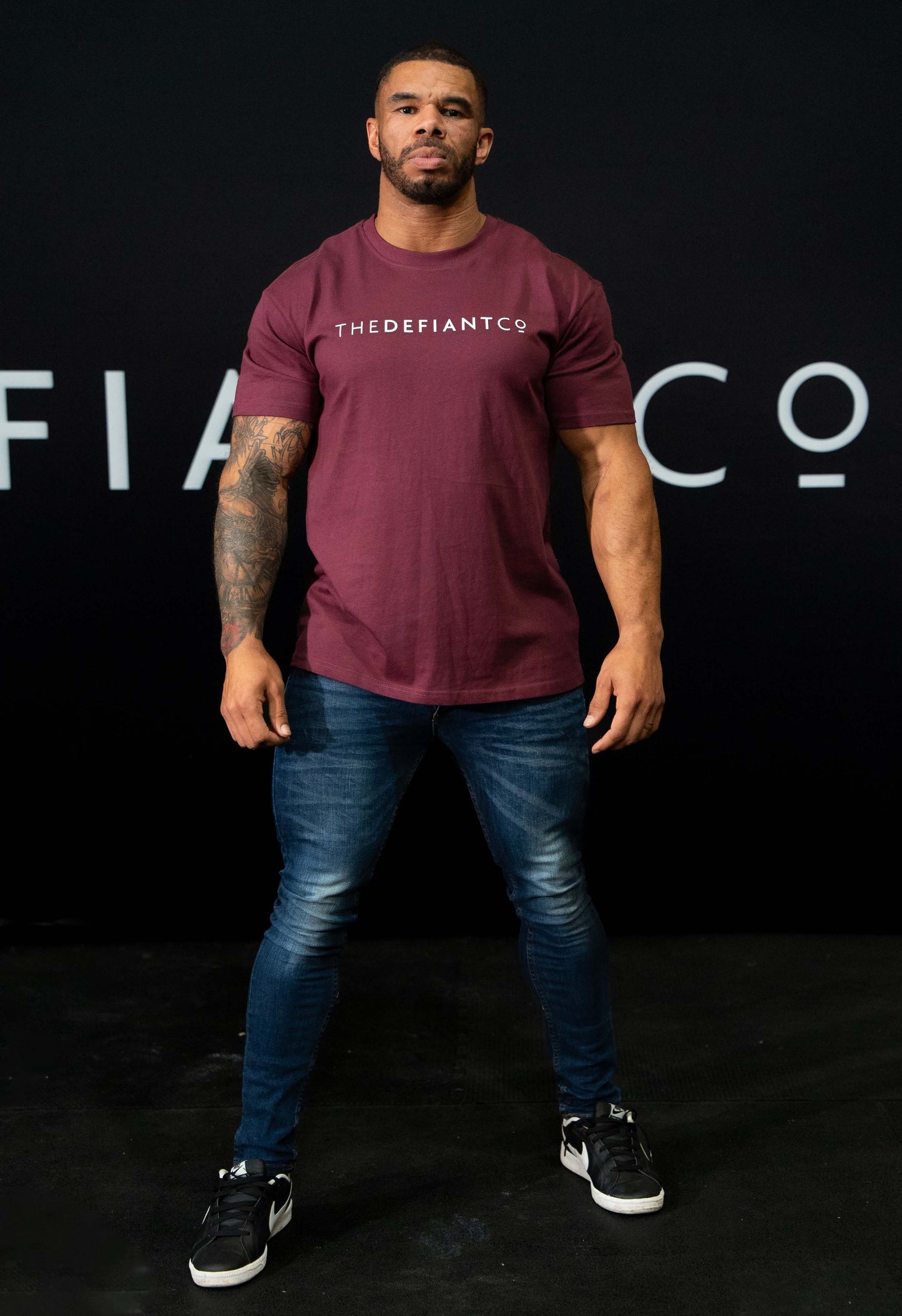 A guy against a black The Defiant Co backdrop wearing the oversized t-shirt.  The tee has the famous 'The Defiant Co' logo boldly displayed across the front. The colour is burgundy.
