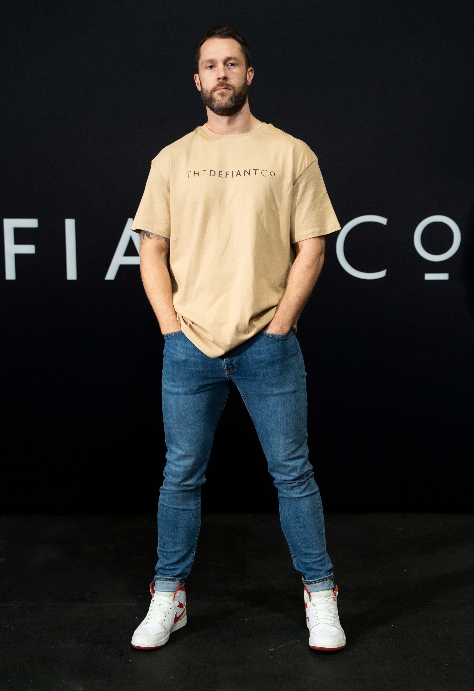 A guy against a black The Defiant Co backdrop wearing the oversized t-shirt.  The tee has the famous 'The Defiant Co' logo boldly displayed across the front. The colour is beige.