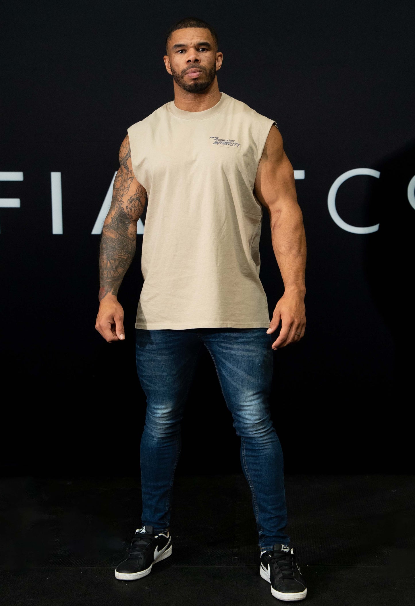 A guy against a black The Defiant Co backdrop wearing the faded khaki proudly refusing to obey authority sleeveless tee and jeans and trainers.