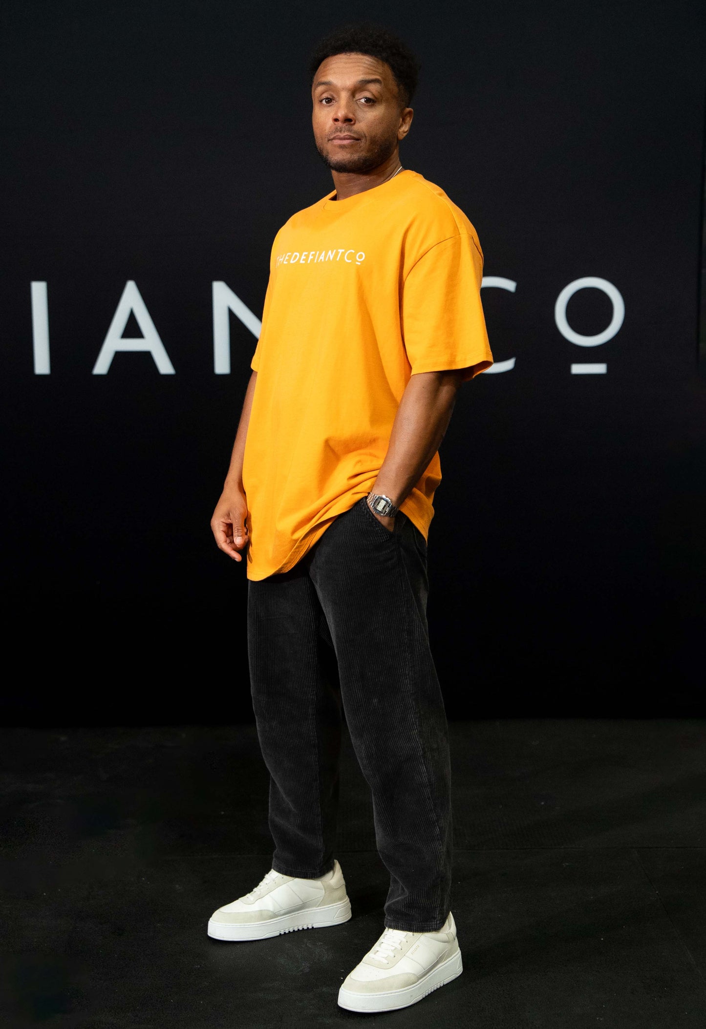 A guy against a black The Defiant Co backdrop wearing the oversized t-shirt.  The tee has the famous 'The Defiant Co' logo boldly displayed across the front. The colour is orange.