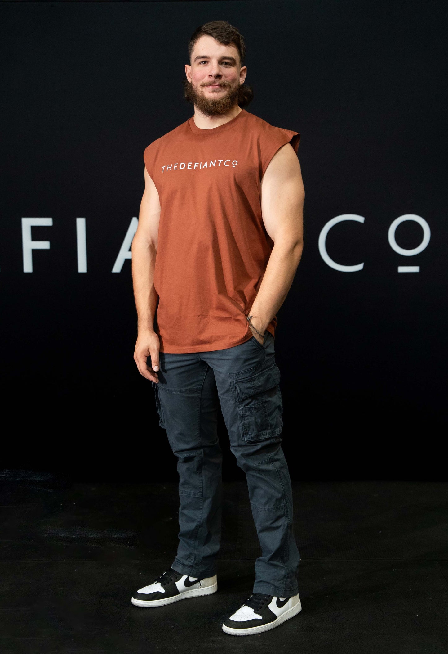 A guy against a black The Defiant Co backdrop wearing the TDCo burnt orange sleeveless tee, with blue trousers and trainers.