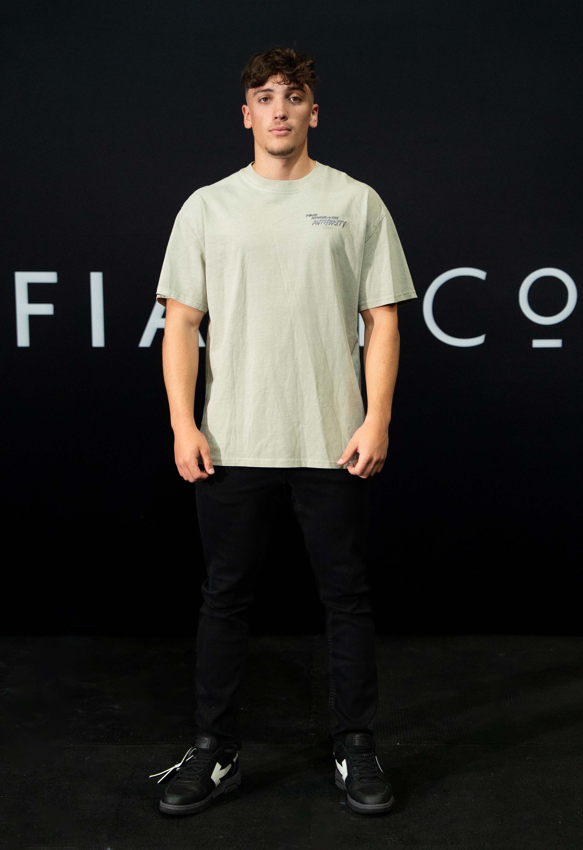 A guy against a black The Defiant Co backdrop wearing the faded khaki proudly refusing to obey authority oversized tee and black jeans.