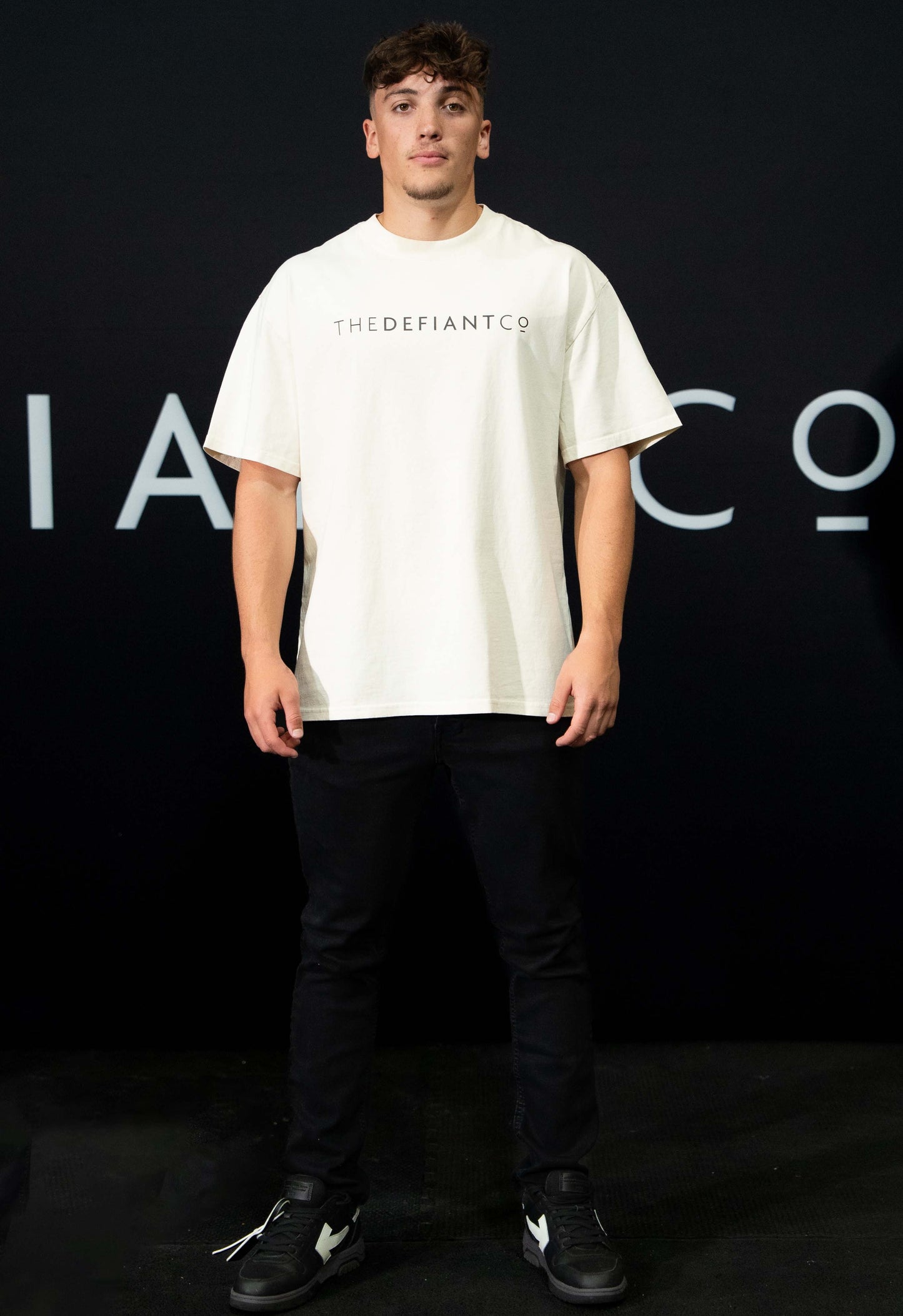 A guy against a black The Defiant Co backdrop wearing the oversized t-shirt.  The tee has the famous 'The Defiant Co' logo boldly displayed across the front. The colour is ecru.
