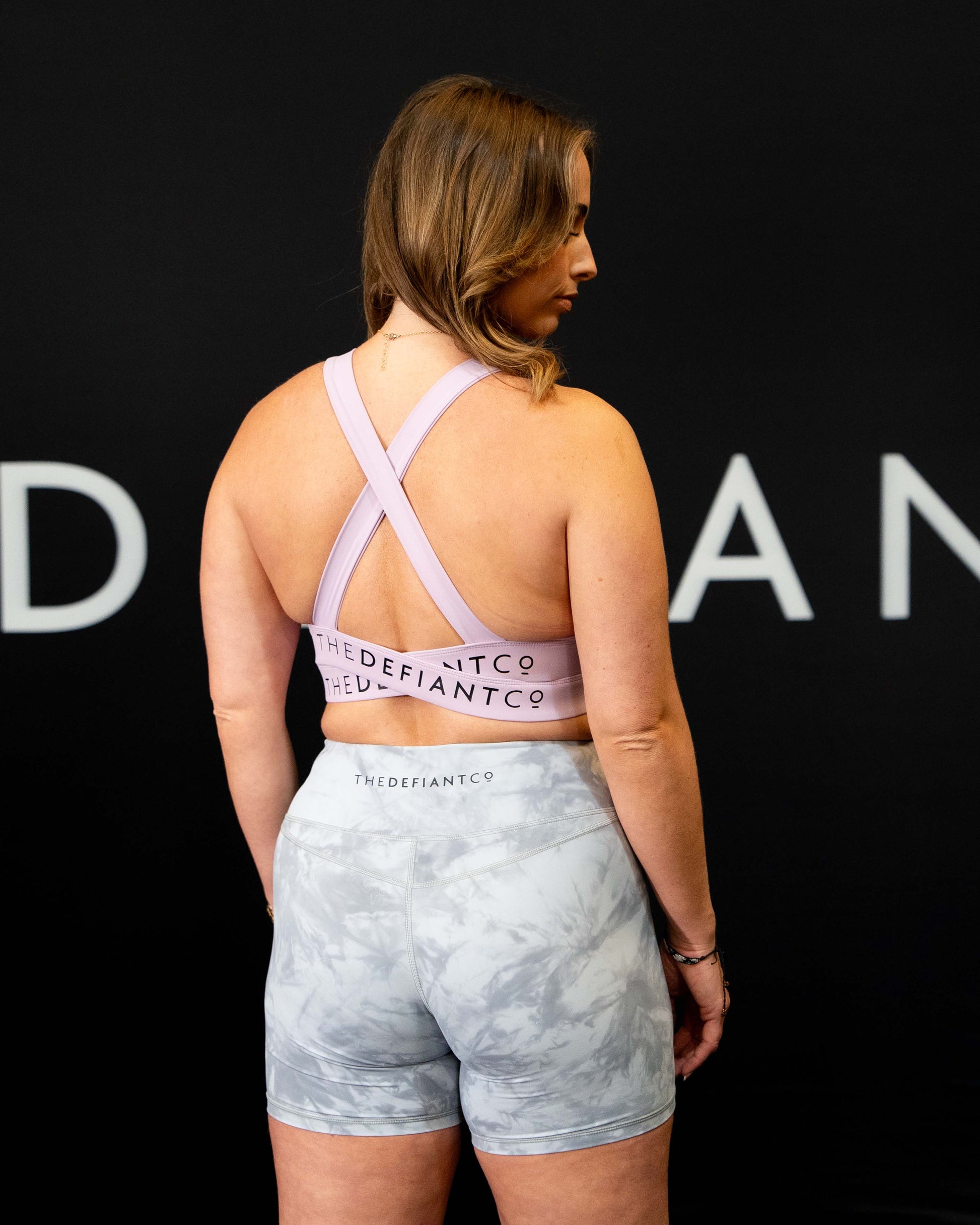 A girl against a black The Defiant Co backdrop wearing the Infinity Sports bra with it's crossed back and supportive front.  The Defiant Co is criss-crossed over both straps across the back. The colour is lilac.