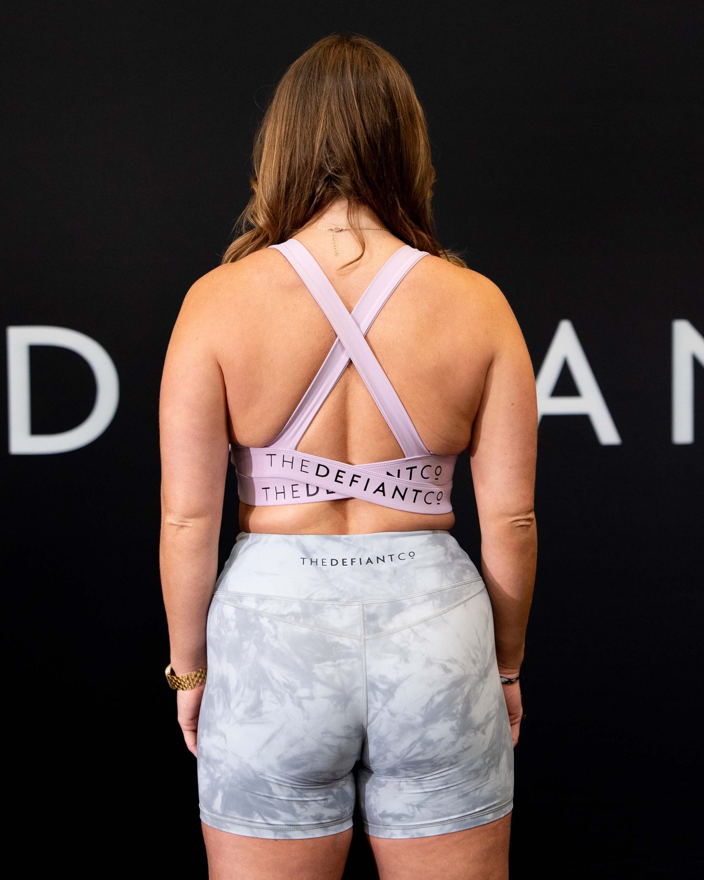 A girl against a black The Defiant Co backdrop wearing the Infinity Sports bra with it's crossed back and supportive front.  The Defiant Co is criss-crossed over both straps across the back. The colour is lilac.