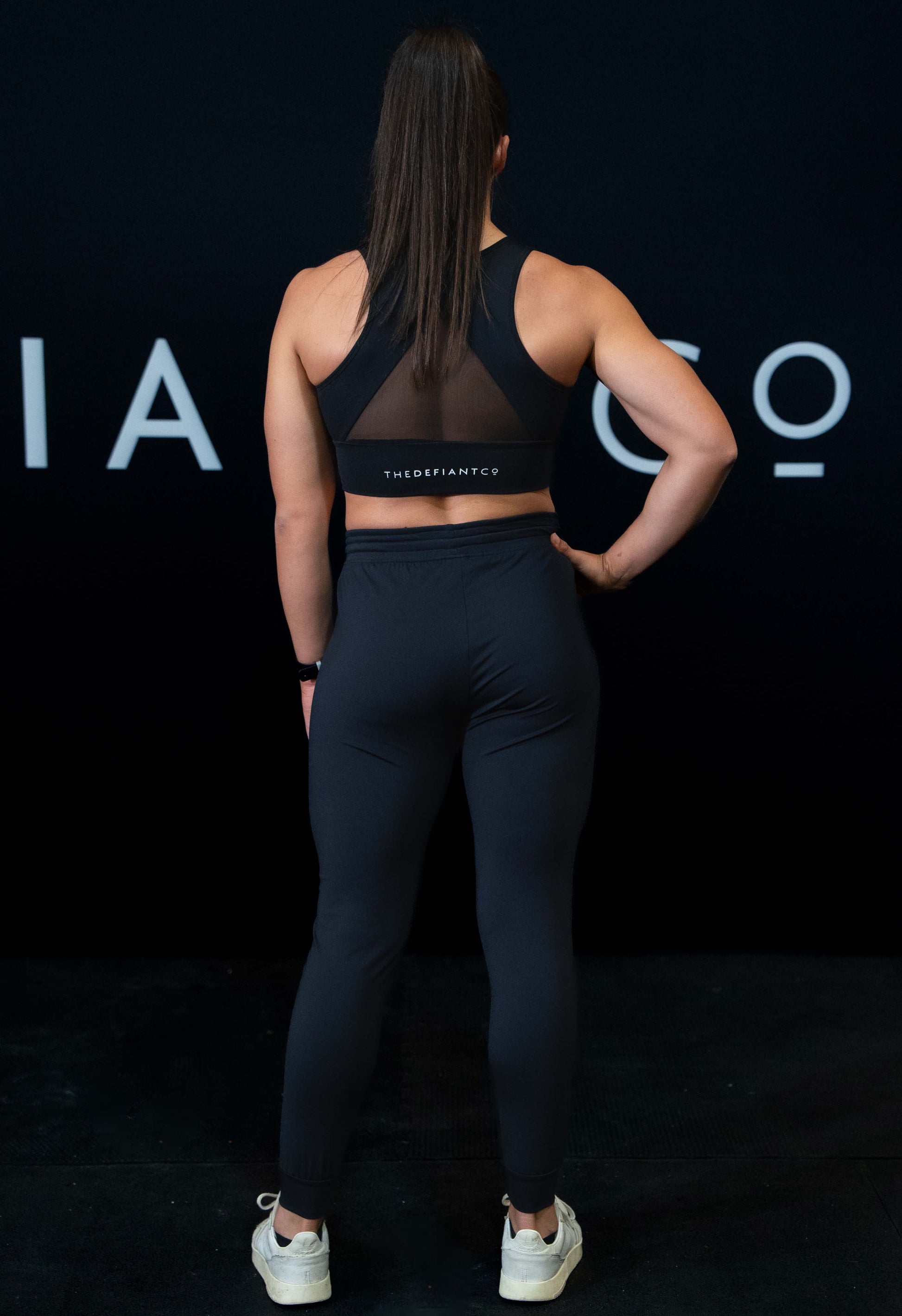 A back of a girl against a black The Defiant Co backdrop wearing the DFNT. Black Jogging Bottoms and black sports bra.   It has a subtle DFNT. logo under the pocket. It has a drawstring waist and the pockets have zips.