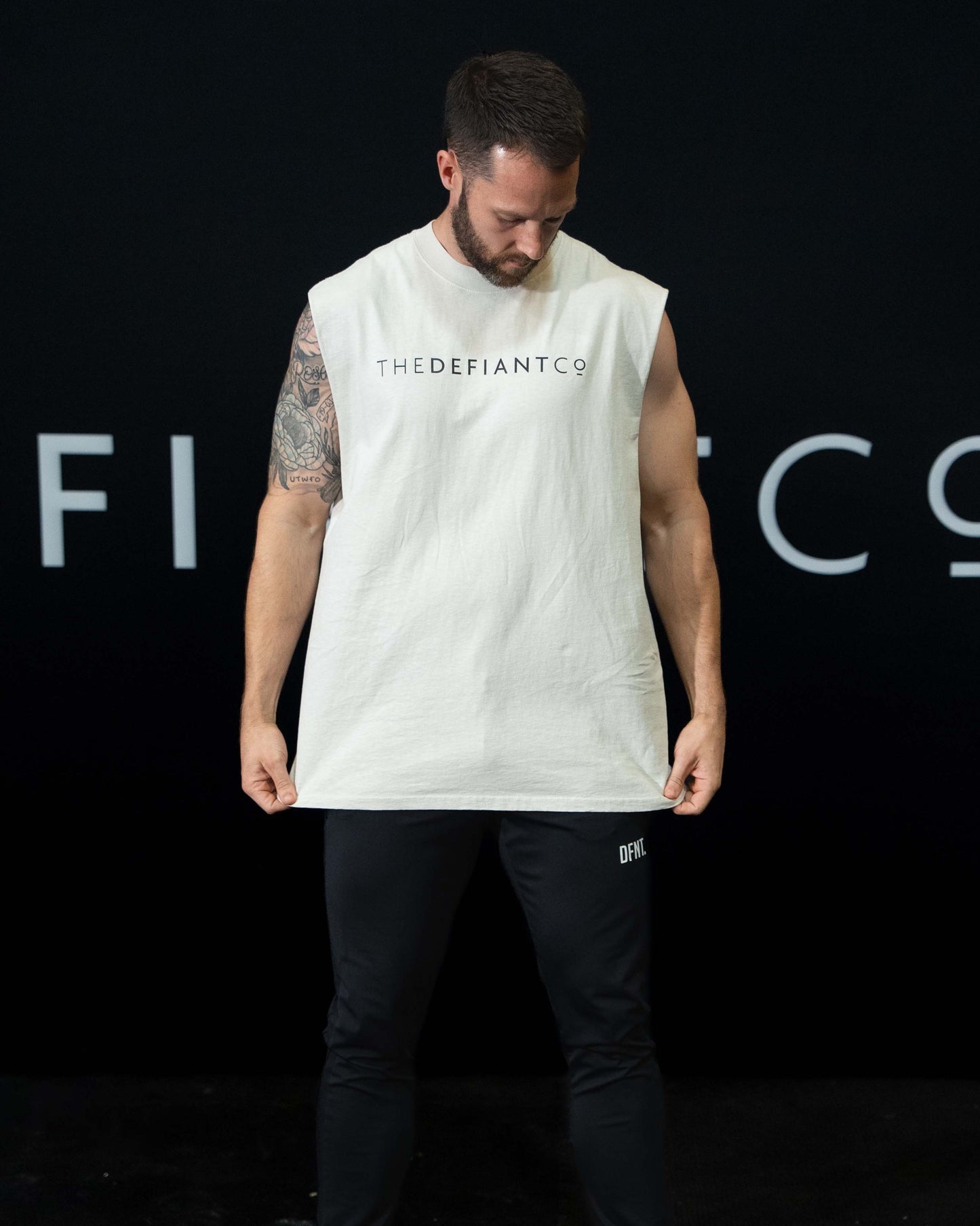 A man against a black The Defiant Co backdrop wearing the DFNT. Black Jogging Bottoms and white sleeveless TDCo tee.   It has a subtle DFNT. logo under the pocket. It has a drawstring waist and the pockets have zips.