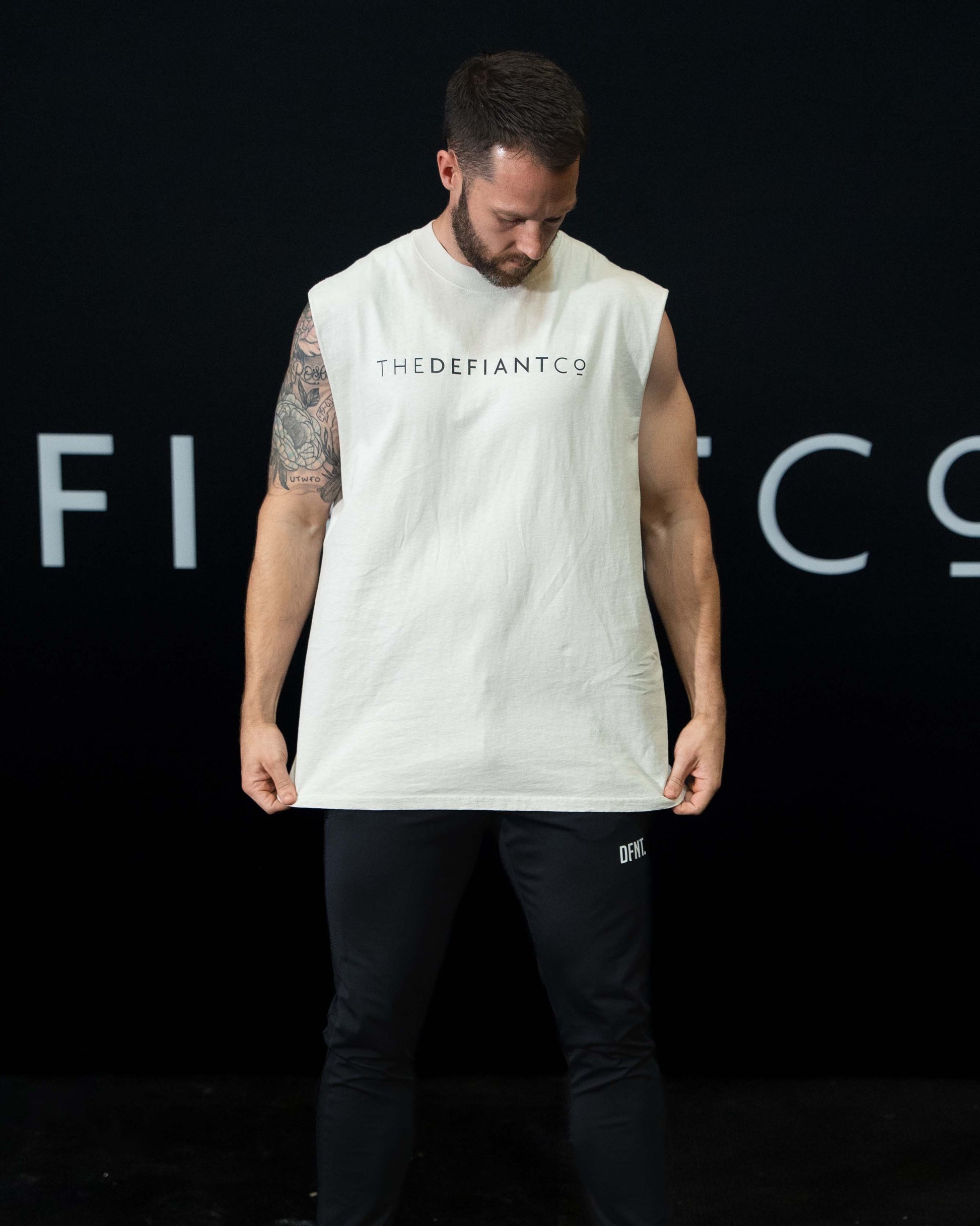 A man against a black The Defiant Co backdrop wearing the DFNT. Black Jogging Bottoms and white sleeveless TDCo tee.   It has a subtle DFNT. logo under the pocket. It has a drawstring waist and the pockets have zips.