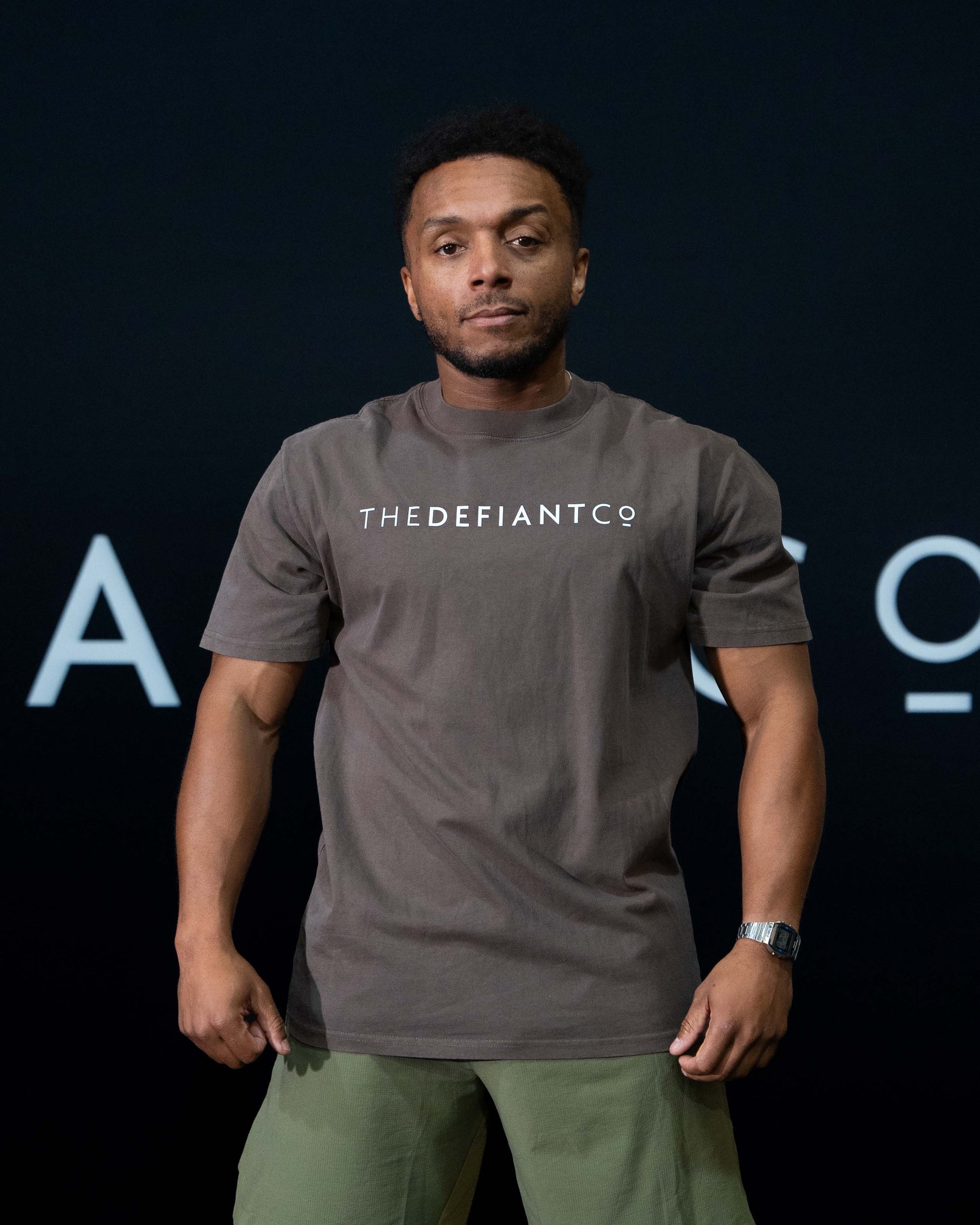A guy against a black The Defiant Co backdrop wearing the oversized t-shirt.  The tee has the famous 'The Defiant Co' logo boldly displayed across the front. The colour is faded brown.