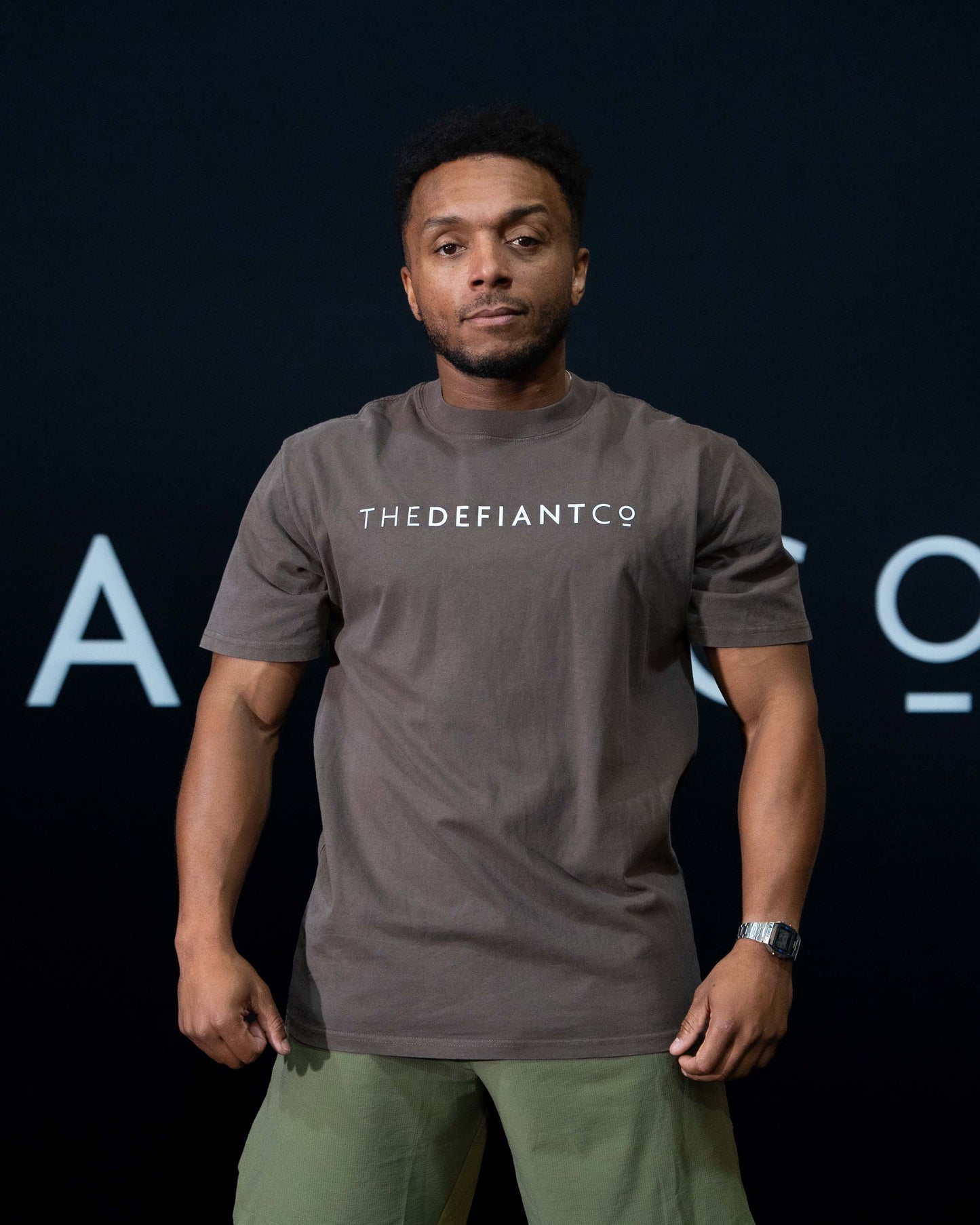 A guy against a black The Defiant Co backdrop wearing the oversized t-shirt. The tee has the famous 'The Defiant Co' logo boldly displayed across the front. The colour is faded brown.