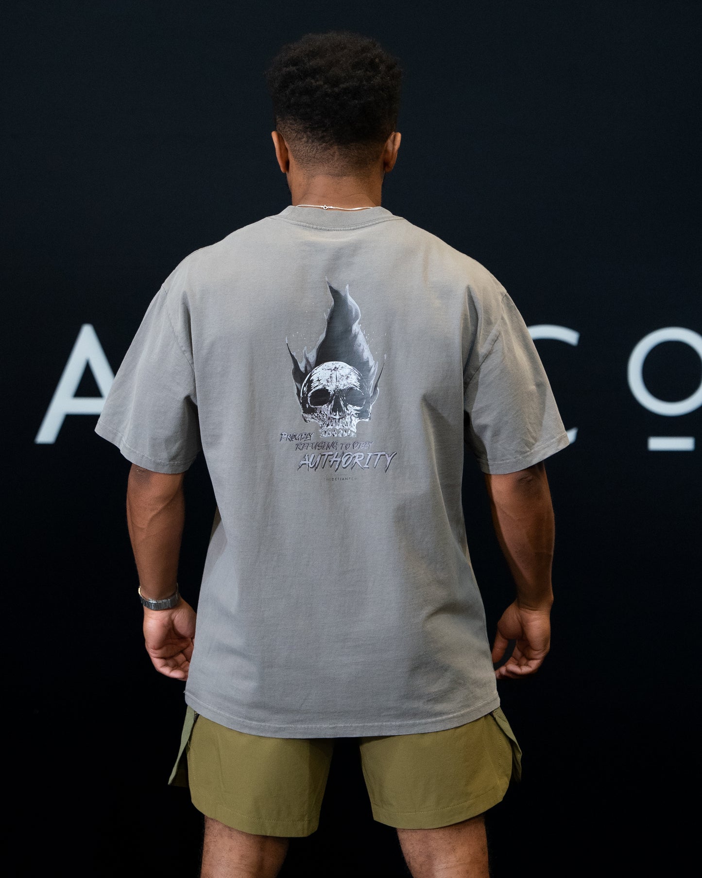 A guy against a black The Defiant Co backdrop wearing the faded grey proudly refusing to obey authority oversized tee and olive green performance shorts.