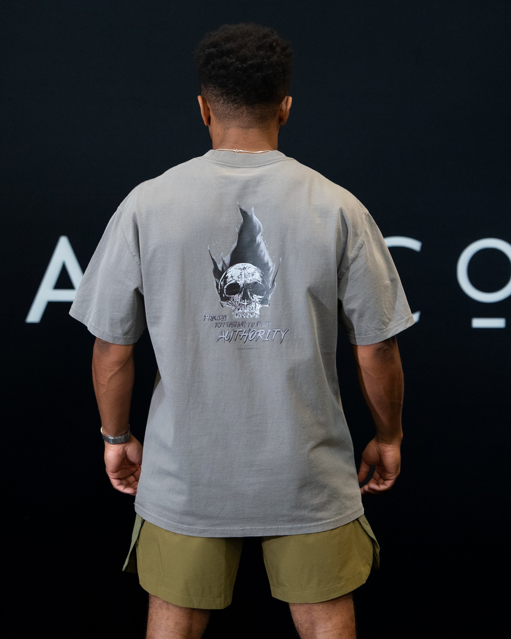 A guy against a black The Defiant Co backdrop wearing the faded grey proudly refusing to obey authority oversized tee and olive green performance shorts.