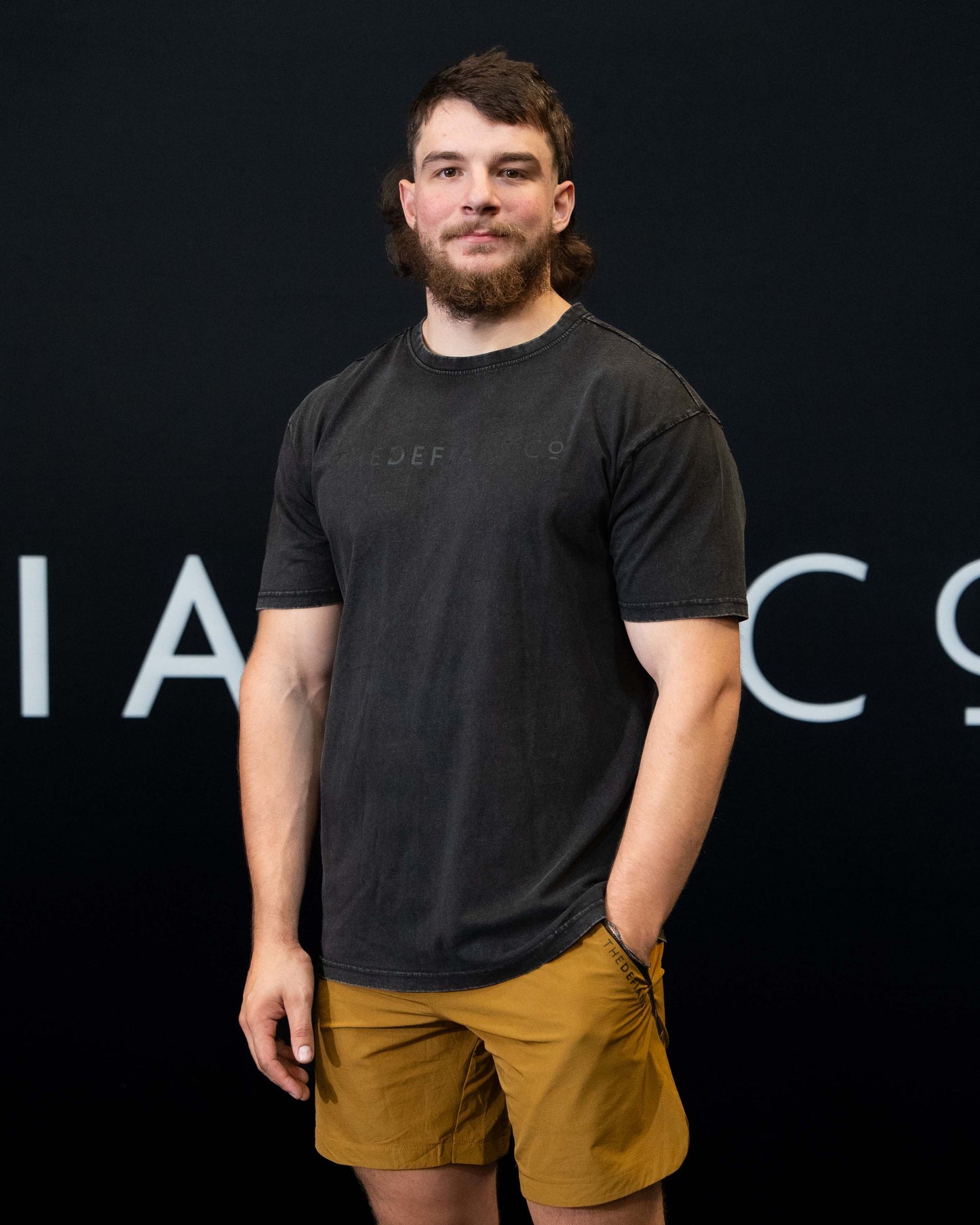 A guy against a black The Defiant Co backdrop wearing the oversized t-shirt.  The tee has the famous 'The Defiant Co' logo boldly displayed across the front. The colour is black on black.