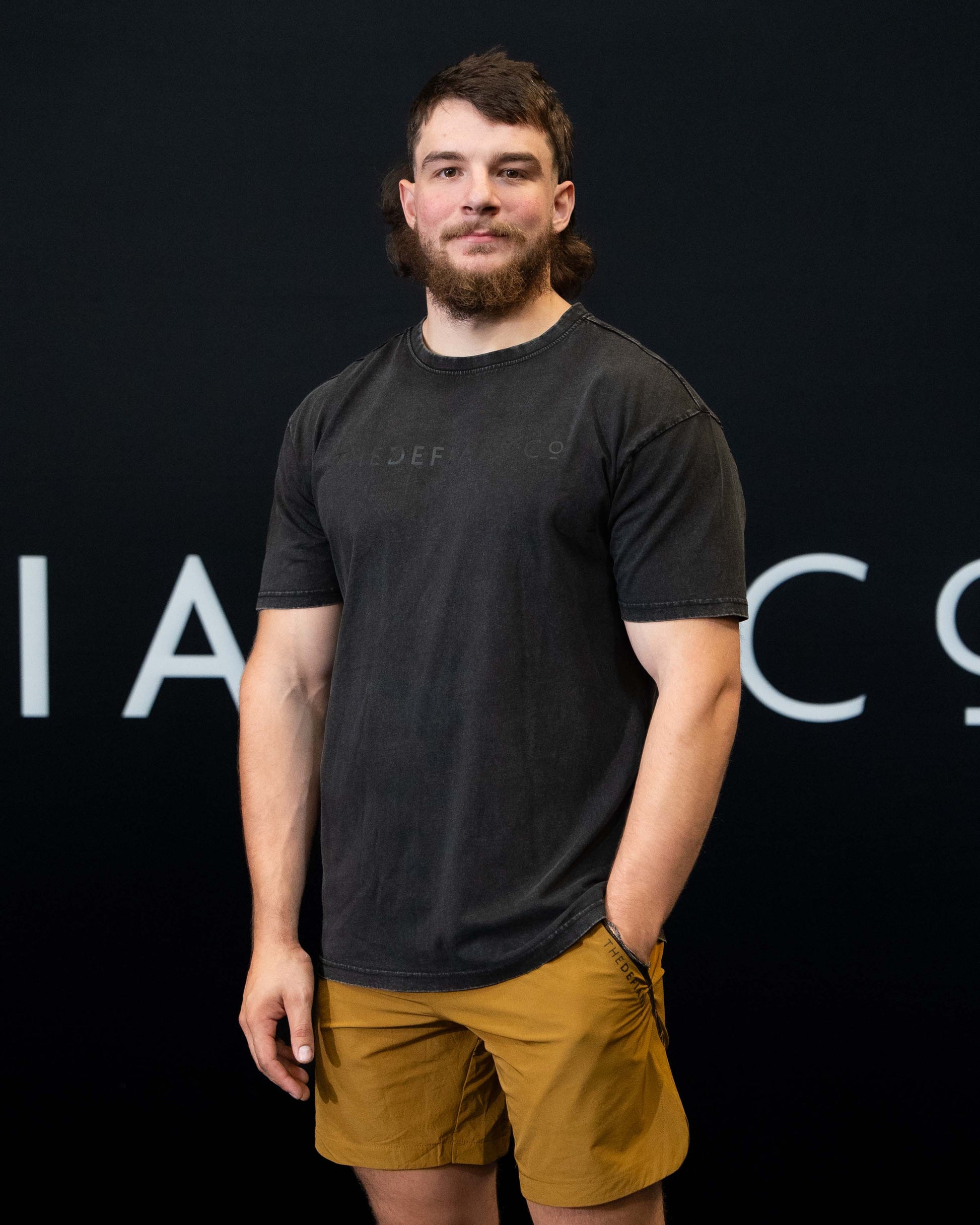 A guy against a black The Defiant Co backdrop wearing the oversized t-shirt.  The tee has the famous 'The Defiant Co' logo boldly displayed across the front. The colour is black on black.