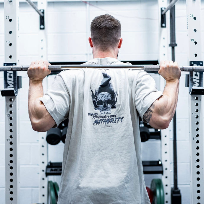 A guy weightlifting wearing the oversized Proudly Refusing To Obey Authority T-Shirt. The guys has big arms and a visible tattoo that matches the defiance of the message on the shirt.