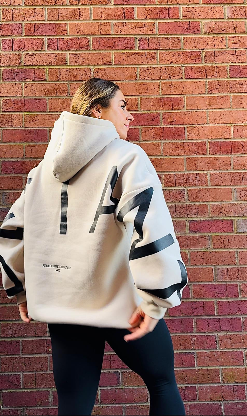 A girl stood in front of some brickwork in an amazing oversized hoodie! The hoodie is off-white and has a bold back DEFIANT print in black that spans both arms and the top of the back. There is also embroidery on the back that reads 'Proudly Refusing To Obey Authority' with 2018 in Roman numerals underneath that. The hoodies are designed to be quite oversized to provide you with that cosy feel.