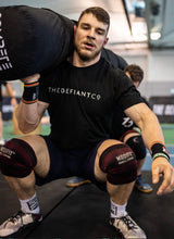 Load image into Gallery viewer, A guy wearing a The Defiant Co standard fit t-shirt whilst performing worm squats during a CrossFit workout at The Defiant Games.  The shirt is black and has the famous The Defiant Co logo across the centre of the chest in white. The shirt is roundneck, 100% cotton and extremely breathable. 