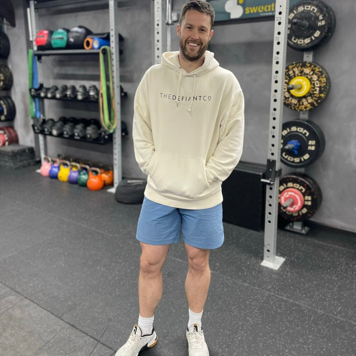 A guy wearing a standard fit, unisex The Defiant Co Hoodie.  The hoodie has draw strings and a pouch pocket on the front as per all standard designs.  The hoodie has the famous The Defiant Co logo across the front of the chest and is plain on the back.  The colour is Sand.