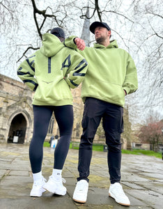 A guy and a girl stood posing in front of a church on a cloudy day, both dressed in the amazing Oversized DFNT. Eternity Hoodies.  The Hoodies have a plain front with a big DEFIANT lettering across the arms and back, a subtle DFNT. tag on the left side and then embroidery stating Proudly Refusing To Obey Authority under the large lettering as well as The Defiant Co embroidered to the left sleeve cuff.  The hoodies are sage green with black logos.