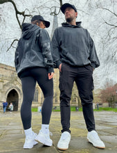 Load image into Gallery viewer, A guy and a girl stood posing in front of a church on a cloudy day, both dressed in the amazing Oversized DFNT. Eternity Hoodies.  The Hoodies have a plain front with a big DEFIANT lettering across the arms and back, a subtle DFNT. tag on the left side and then embroidery stating Proudly Refusing To Obey Authority under the large lettering as well as The Defiant Co embroidered to the left sleeve cuff.  The hoodies are washed black with black logos.