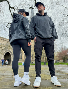 A guy and a girl stood posing in front of a church on a cloudy day, both dressed in the amazing Oversized DFNT. Eternity Hoodies.  The Hoodies have a plain front with a big DEFIANT lettering across the arms and back, a subtle DFNT. tag on the left side and then embroidery stating Proudly Refusing To Obey Authority under the large lettering as well as The Defiant Co embroidered to the left sleeve cuff.  The hoodies are washed black with black logos.