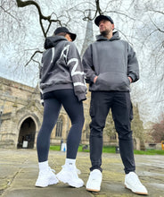 Load image into Gallery viewer, A guy and a girl stood posing in front of a church on a cloudy day, both dressed in the amazing Oversized DFNT. Eternity Hoodies.  The Hoodies have a plain front with a big DEFIANT lettering across the arms and back, a subtle DFNT. tag on the left side and then embroidery stating Proudly Refusing To Obey Authority under the large lettering as well as The Defiant Co embroidered to the left sleeve cuff.  The hoodies are charcoal with white logos.