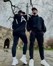 Load image into Gallery viewer, A guy and a girl stood posing in front of a church on a cloudy day, both dressed in the amazing Oversized DFNT. Eternity Hoodies.  The Hoodies have a plain front with a big DEFIANT lettering across the arms and back, a subtle DFNT. tag on the left side and then embroidery stating Proudly Refusing To Obey Authority under the large lettering as well as The Defiant Co embroidered to the left sleeve cuff.  The hoodies are black with white logos.