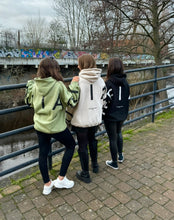 Load image into Gallery viewer, Three girls on a bridge overlooking a canal in amazing oversized hoodies! The hoodies are sage green, off-white and black respectively and have a bold back DEFIANT print that spans both arms and the top of the back.  There is also embroidery on the back that reads &#39;Proudly Refusing To Obey Authority&#39; with 2018 in Roman numerals underneath that.  The hoodies are designed to be quite oversized to provide you with that cosy feel.