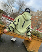 Load image into Gallery viewer, A sat on a park bench in Sheffield in an amazing oversized hoodie! The hoodie is sage green and has a bold back DEFIANT print in black that spans both arms and the top of the back. There is also embroidery on the back that reads &#39;Proudly Refusing To Obey Authority&#39; with 2018 in Roman numerals underneath that. The hoodies are designed to be quite oversized to provide you with that cosy feel.