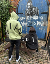 Load image into Gallery viewer, A guy and girl stood infront of some awesome artwork that embodies the Defiant spirit in amazing oversized hoodies! The hoodies are sage green and black and have a bold back DEFIANT print that spans both arms and the top of the back. There is also embroidery on the back that reads &#39;Proudly Refusing To Obey Authority&#39; with 2018 in Roman numerals underneath that. The hoodies are designed to be quite oversized to provide you with that cosy feel.