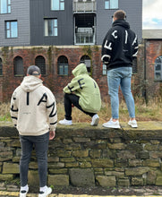 Load image into Gallery viewer, Three guys on a bridge overlooking a canal in amazing oversized hoodies! The hoodies are sage green, off-white and black respectively and have a bold back DEFIANT print that spans both arms and the top of the back. There is also embroidery on the back that reads &#39;Proudly Refusing To Obey Authority&#39; with 2018 in Roman numerals underneath that. The hoodies are designed to be quite oversized to provide you with that cosy feel.